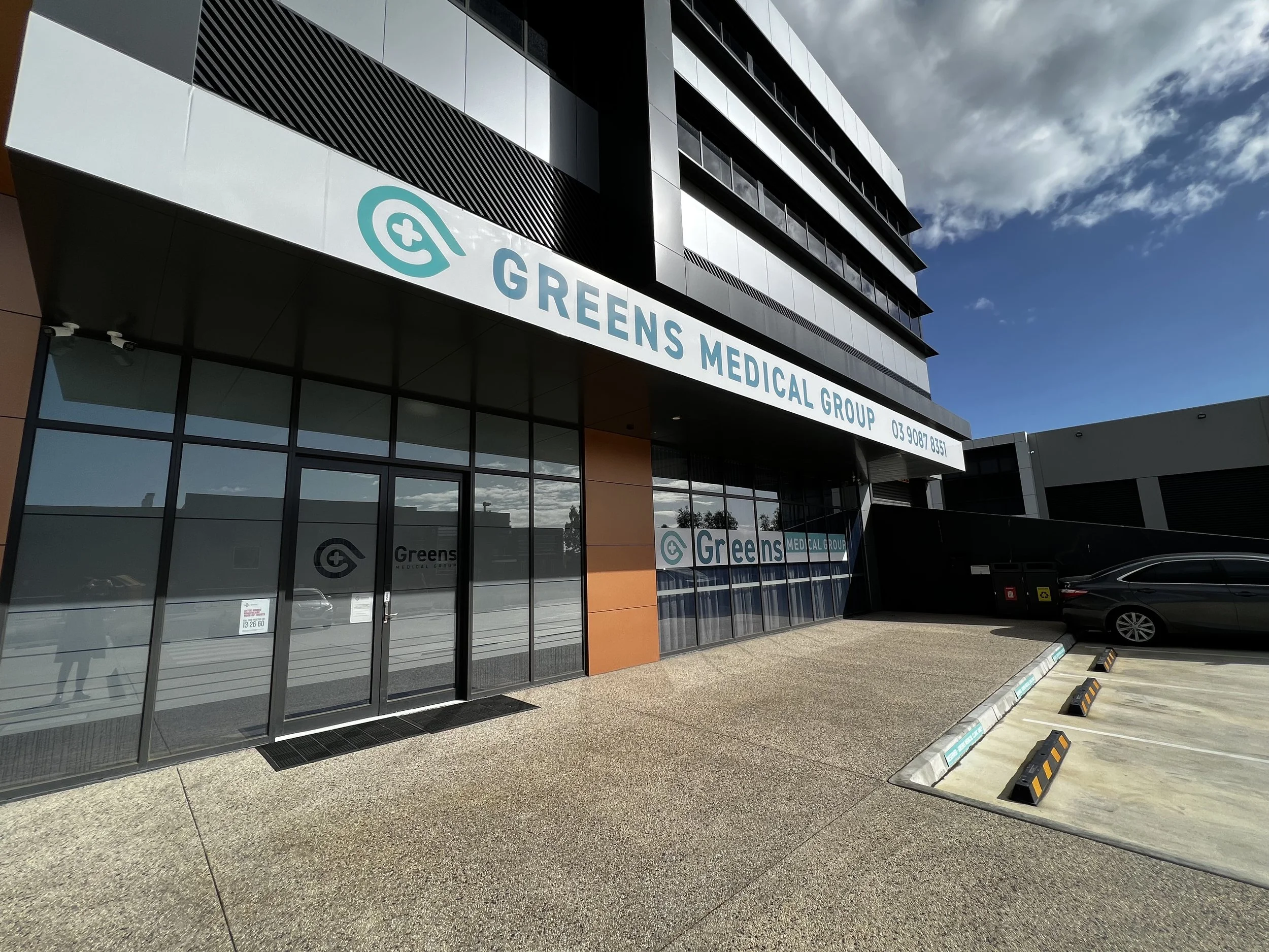 Exterior view of Greens Medical Group building with glass doors and windows, parking lot with a few cars, sign with the clinic's name, and partly cloudy sky.