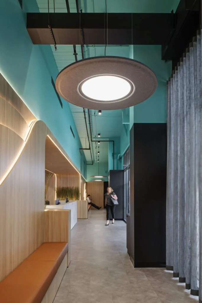 Modern interior with teal walls, large circular ceiling light, wooden paneling with built-in lighting, a cushioned bench, and people walking and sitting.