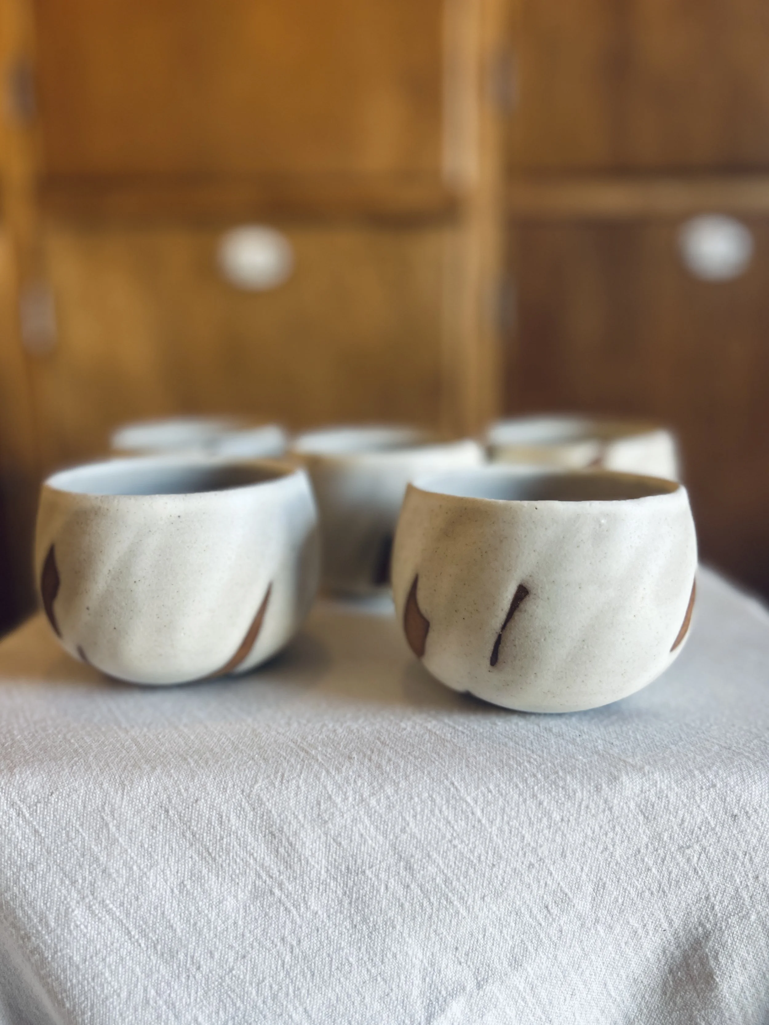 Six small white ceramic cups with dark brown decorative lines, placed on a white textured surface with a wooden cabinet in the background.