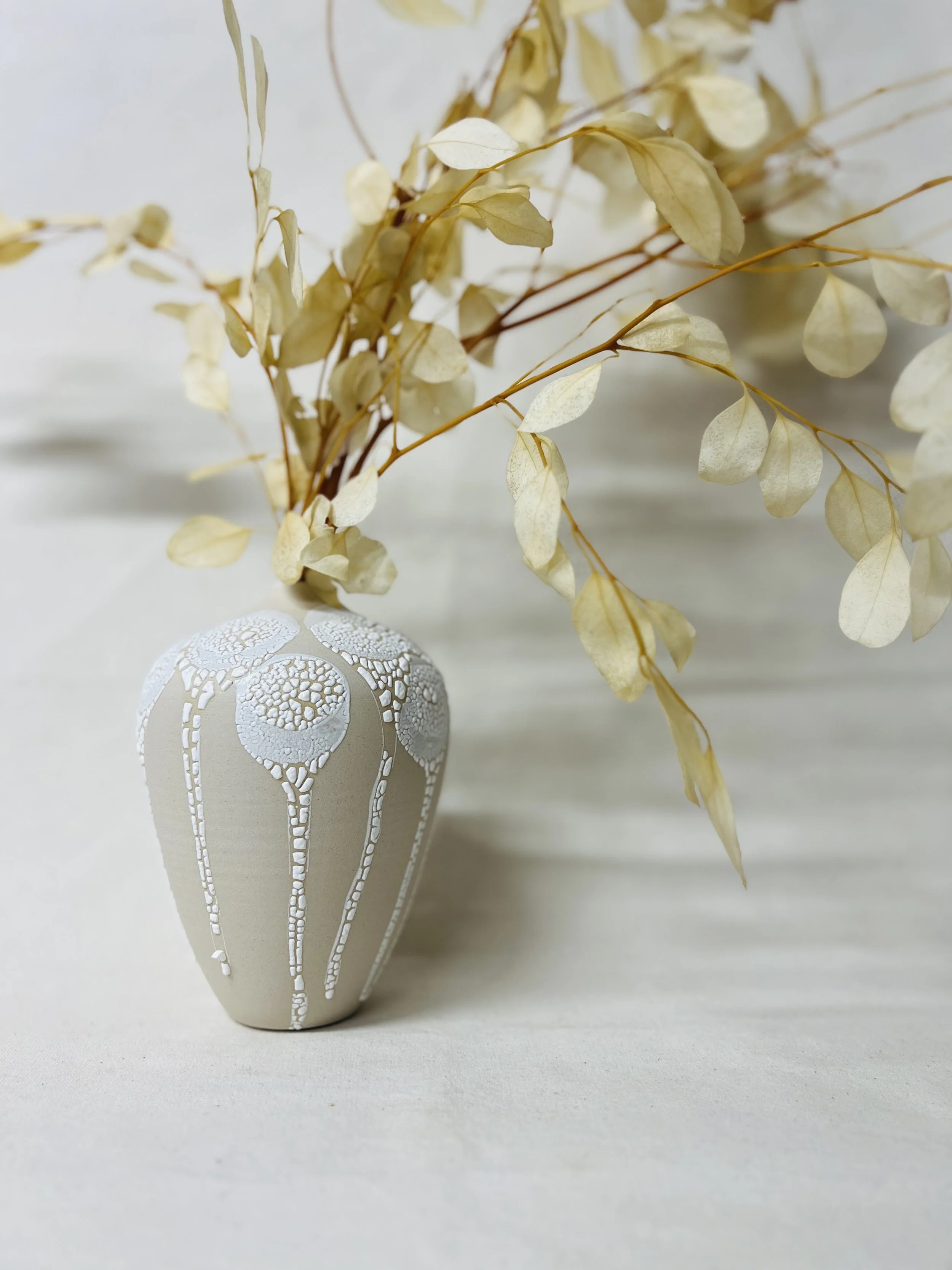 A beige decorative vase with white leaf patterns, holding dried cream-colored leaves with thin stems, resting on a light-colored surface against a white background.