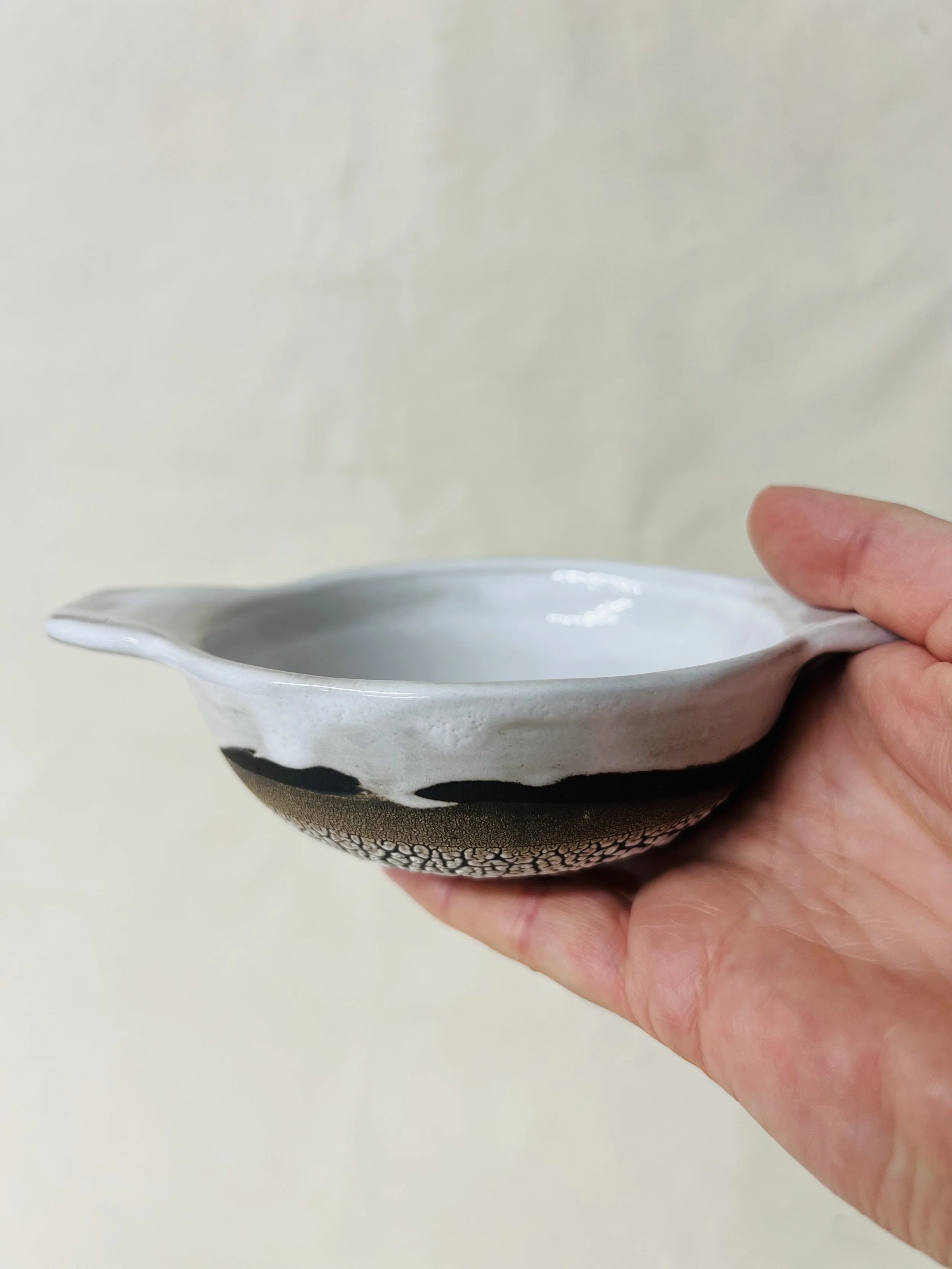 A hand holding a small white and black ceramic dish with a textured black pattern around the bottom rim, against a plain light-colored background.