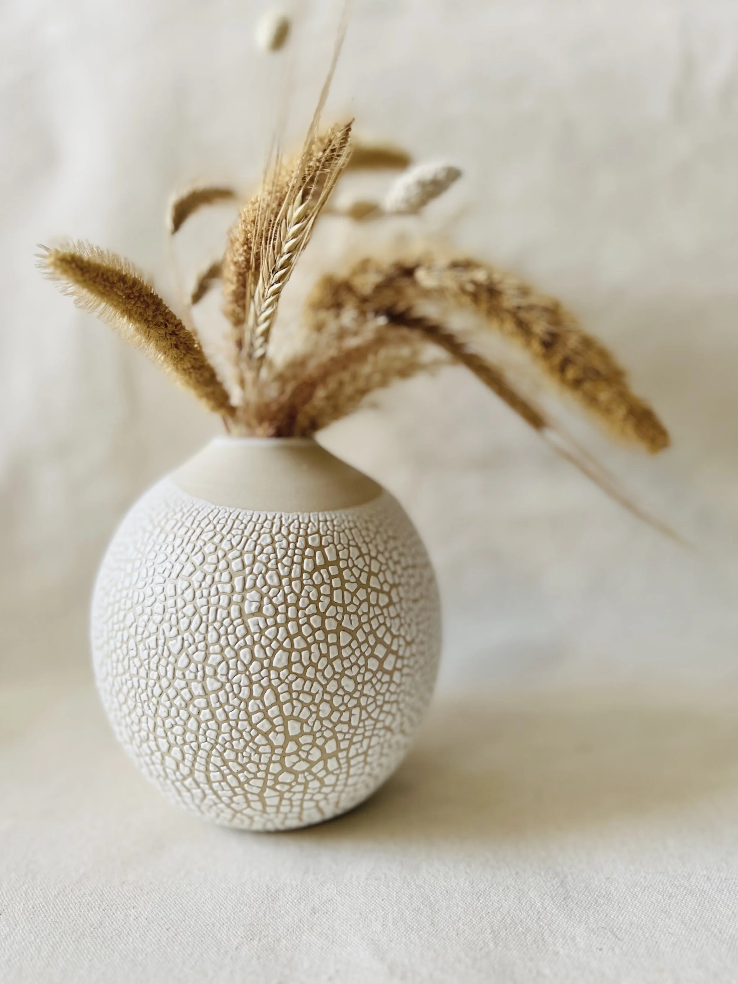 A white textured ceramic vase with a crackled pattern, holding dried grasses and wheat stalks. The background is a plain, light-colored surface.