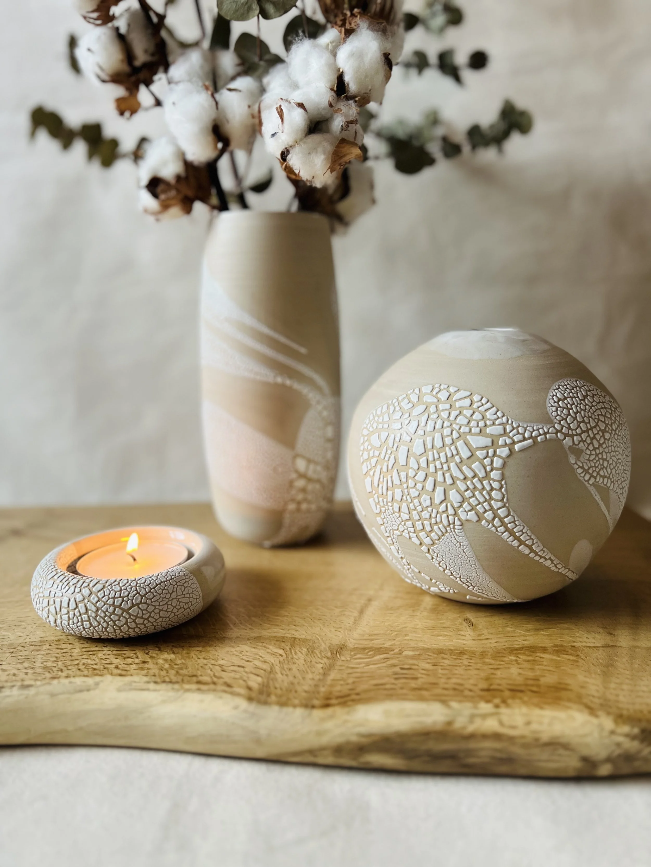 A beige ceramic vase with a cotton branch and eucalyptus leaves, a round porcelain object with a crackle pattern, and a lit candle on a wooden tray.