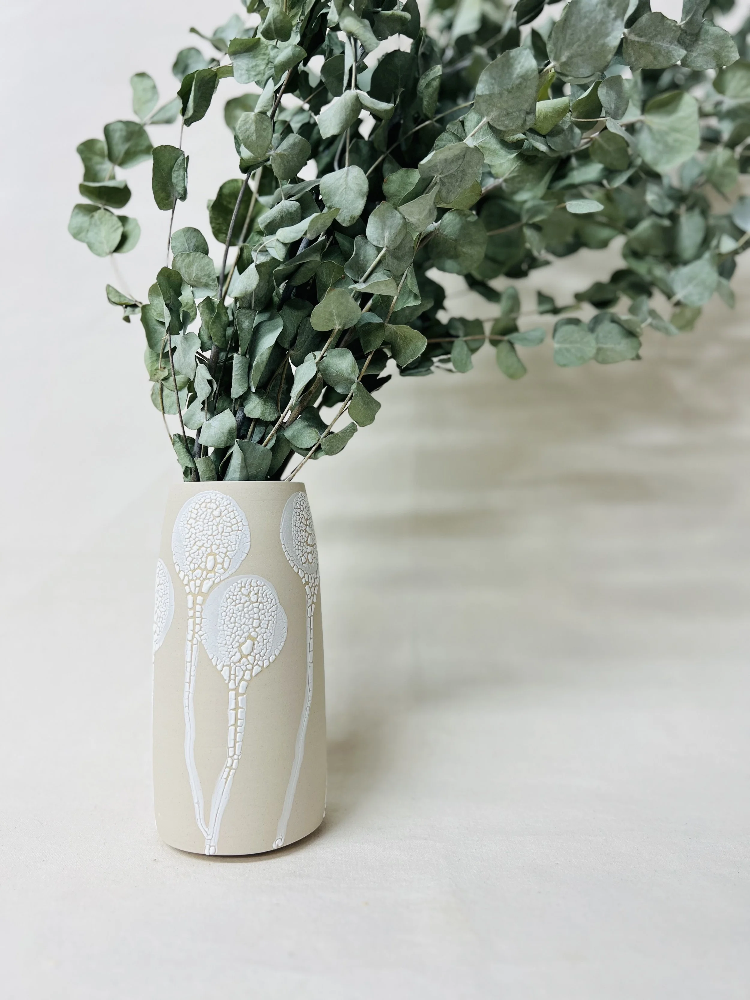 A beige ceramic vase with white abstract tree patterns, holding a bunch of green leafy foliage, placed on a plain surface against a light background.