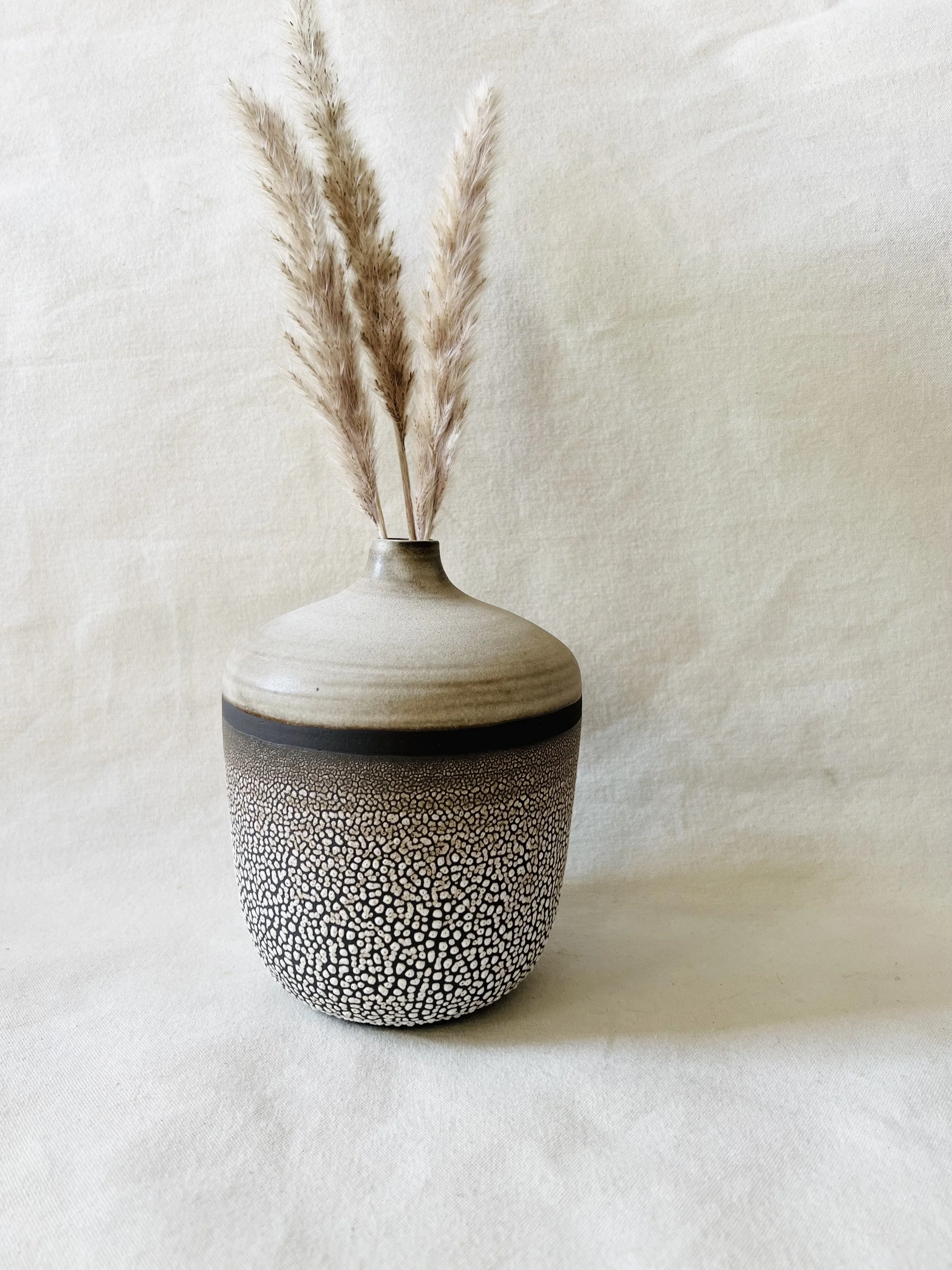A modern ceramic vase with a textured black and white pattern, containing tall beige pampas grass, placed against a plain beige background.