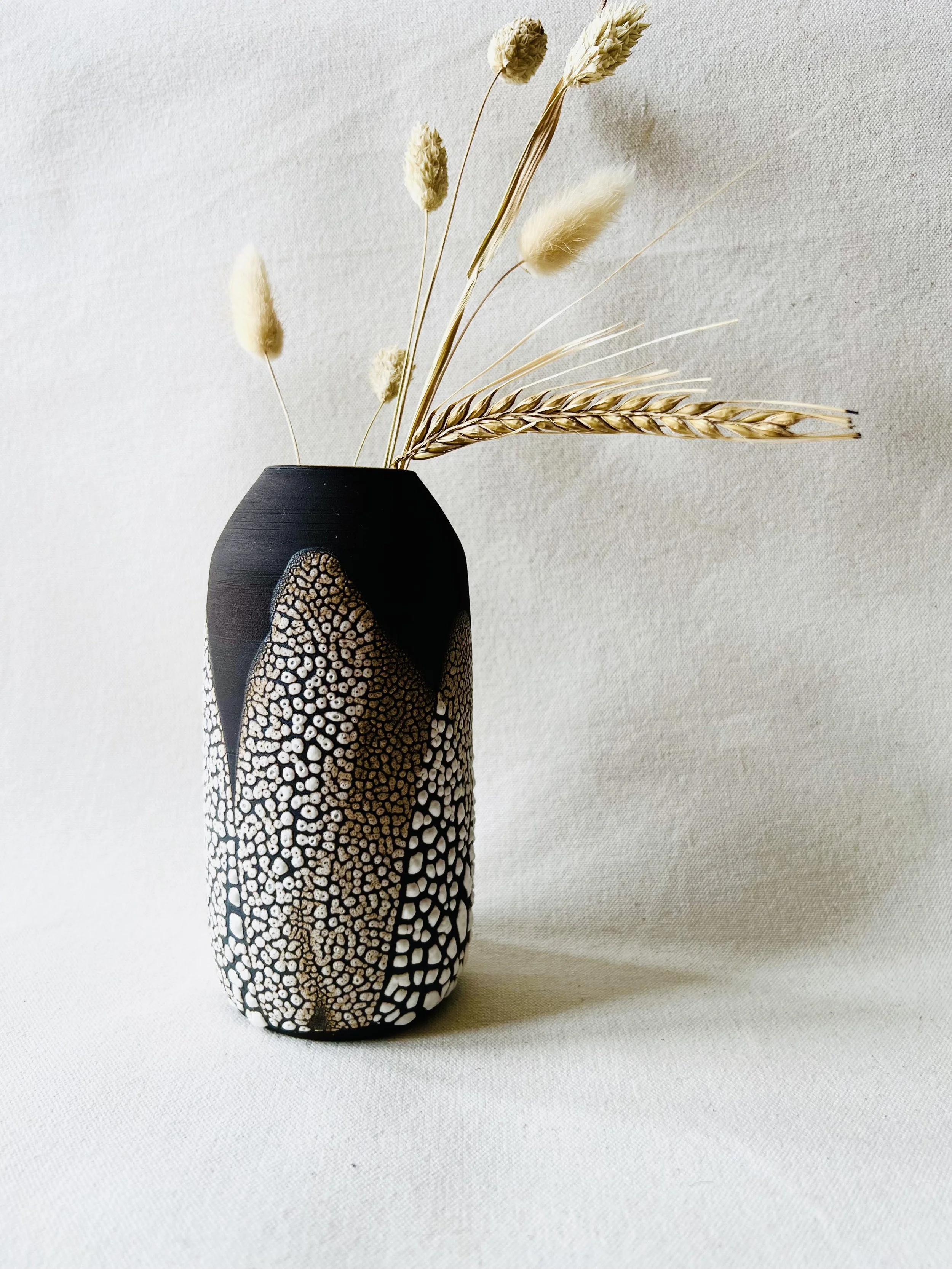 A black vase with white and brown patterned design, holding dried beige and light brown grasses and seed heads, against a plain light background.
