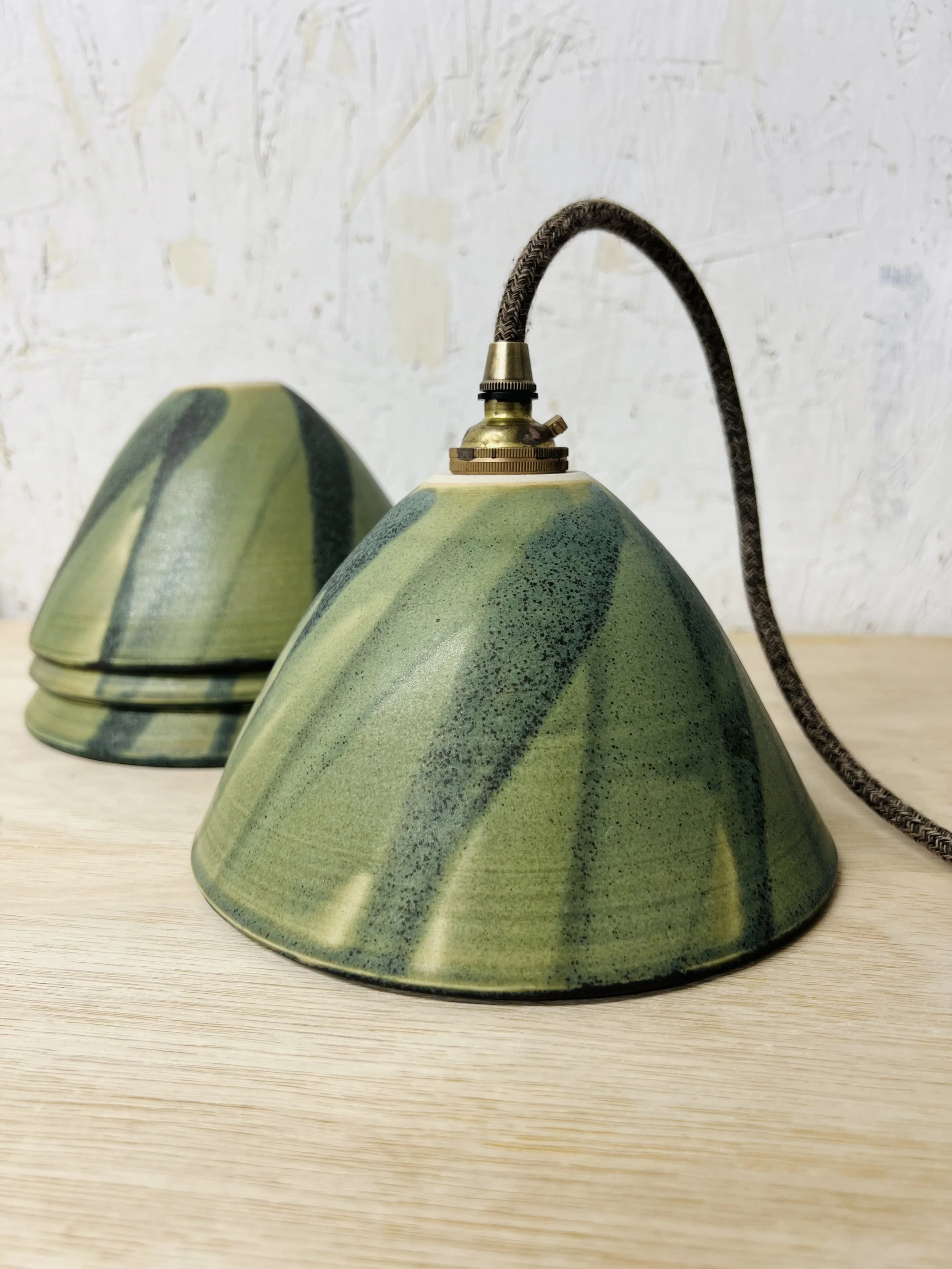 Close-up of a green ceramic table lamp with a black cord, on a light wooden surface, against a textured light-colored wall.