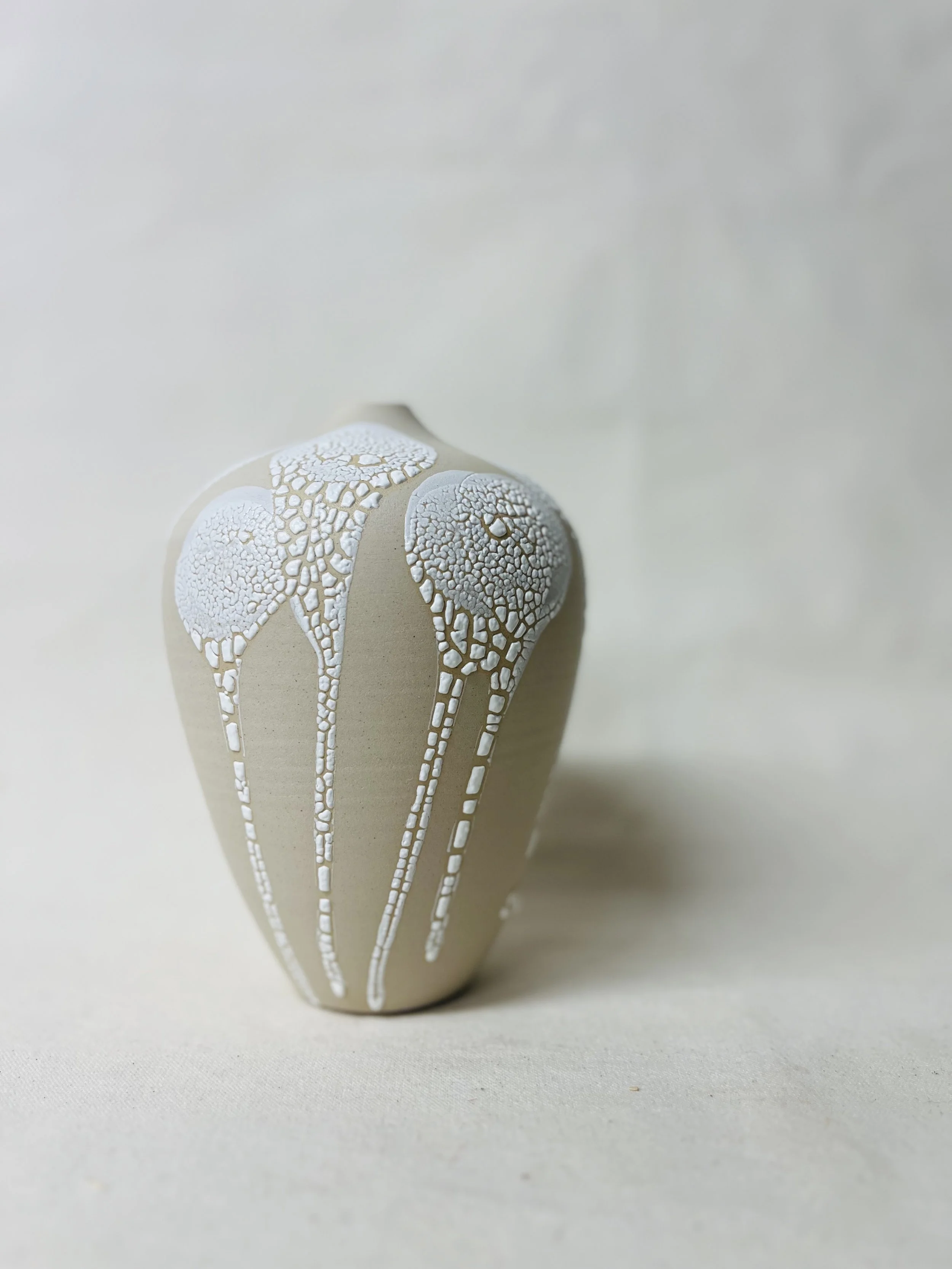 A beige ceramic vase with white textured floral and leaf patterns raised on its surface, set against a plain, light background.