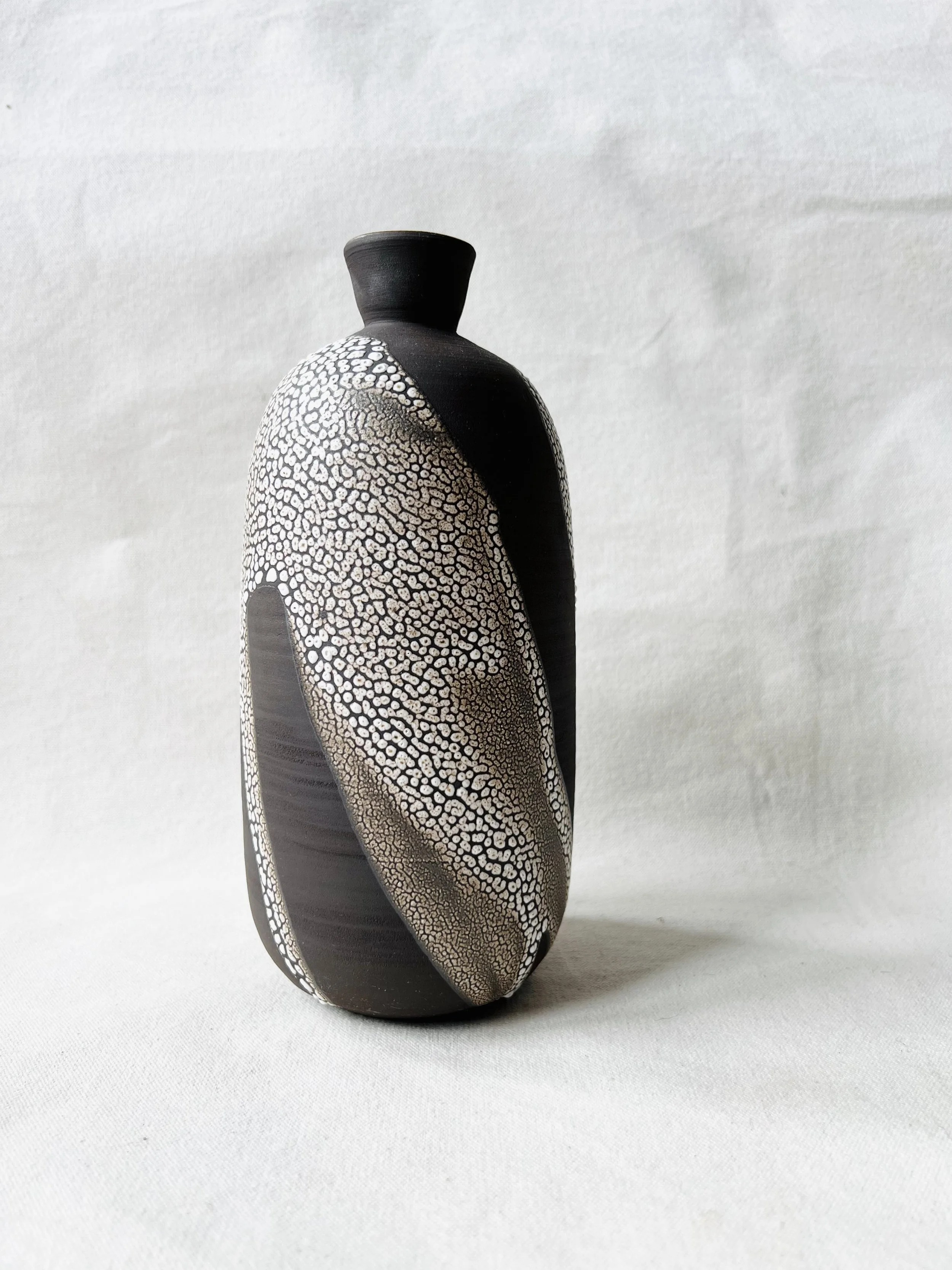 A black and white ceramic vase with a textured pattern resembling small bubbles or scales, and a slender neck, placed on a plain white surface with a plain white background.