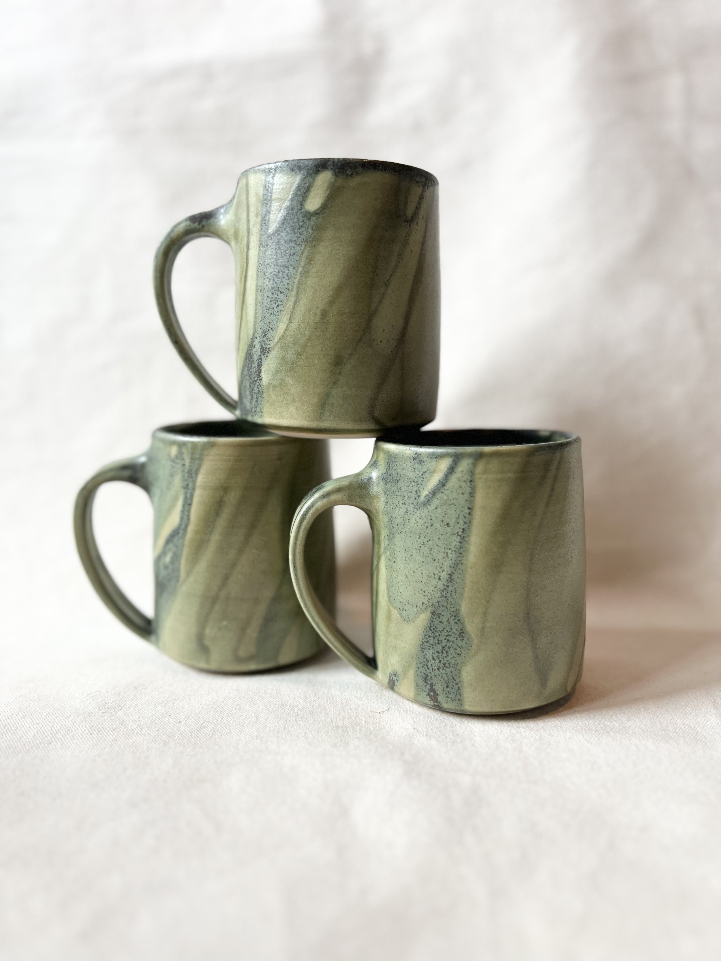 Three ceramic mugs with green marbled glaze, stacked on a white surface against a plain light background.