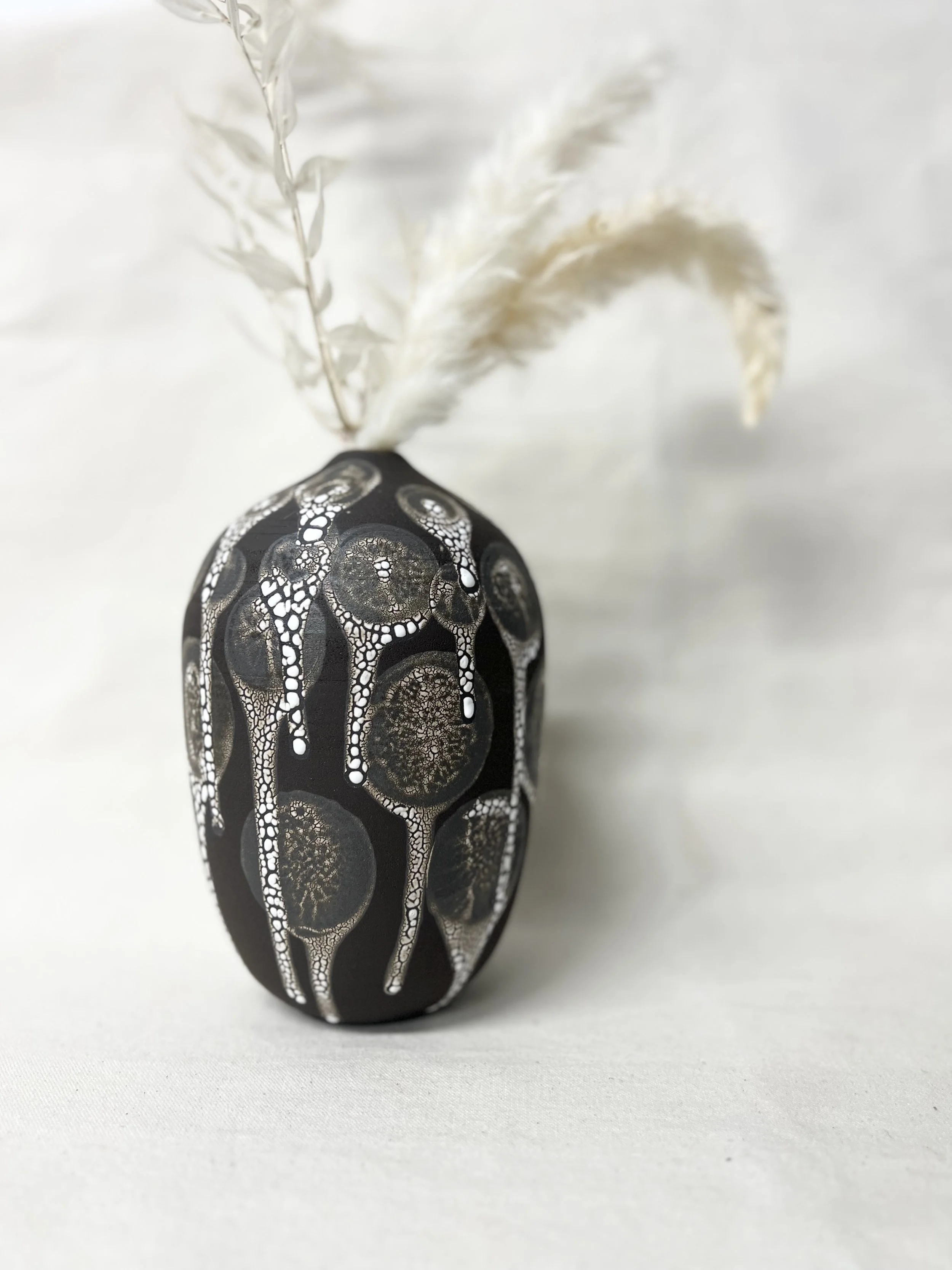 A black eggshell-shaped vase with white intricate dot patterns, holding white feathered plant stems, placed on a white surface with a plain white background.