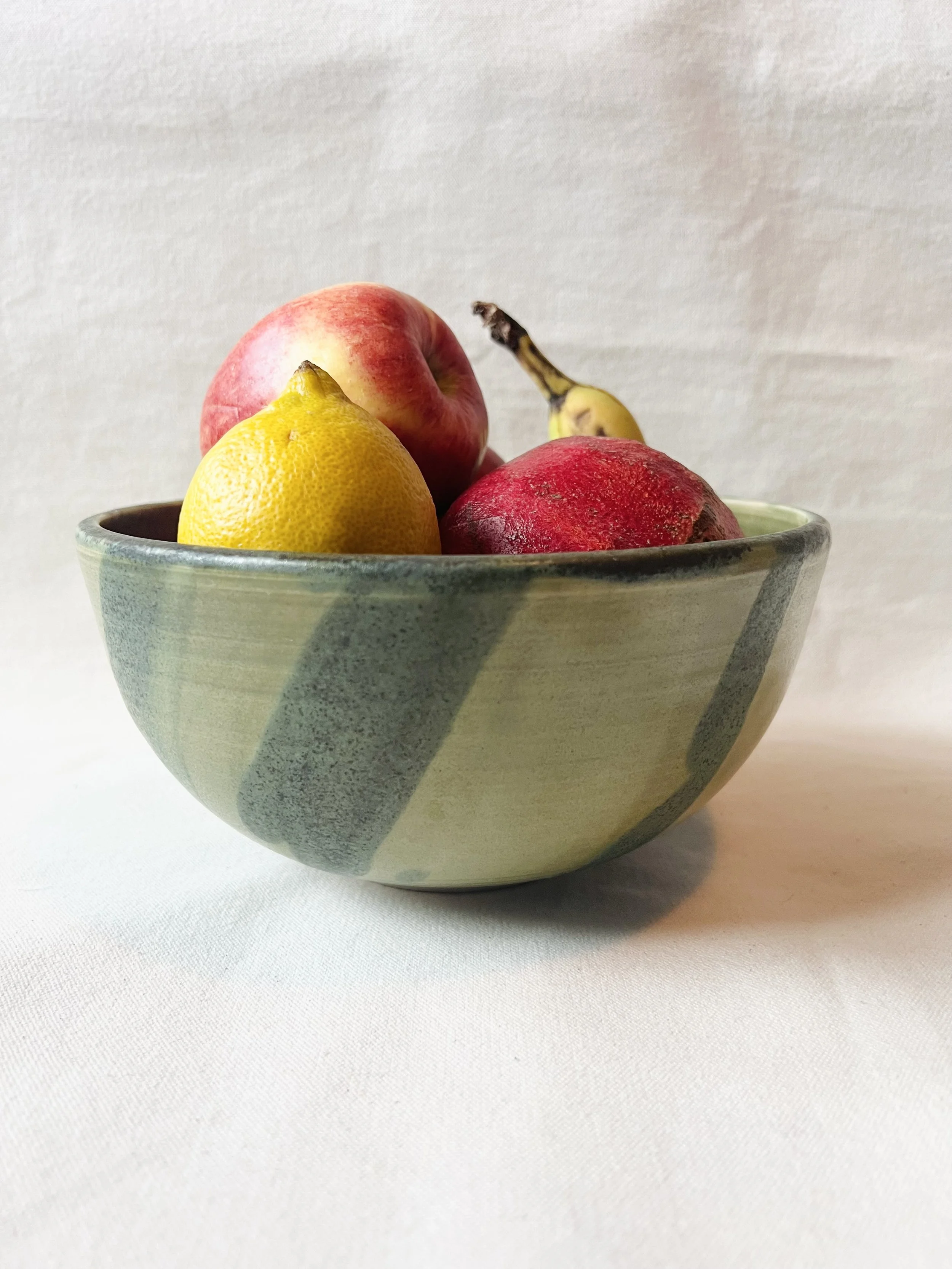 A ceramic bowl with a speckled gray and white pattern, containing a yellow lemon, a red apple, a red pomegranate, and a small banana with a brown stem against a plain light background.