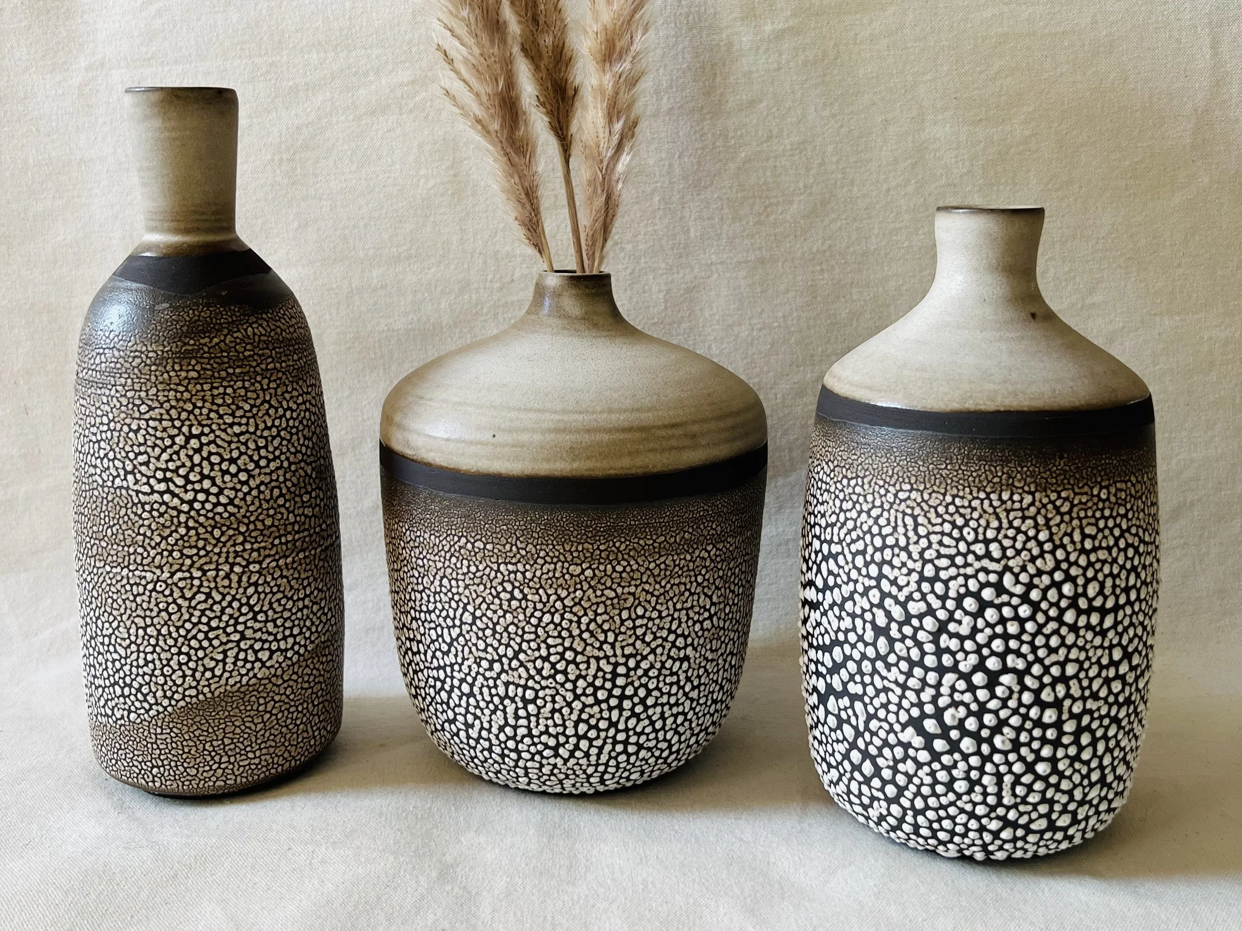 Three decorative vases with textured black and beige designs, one contains dried pampas grass, set against a plain beige background.