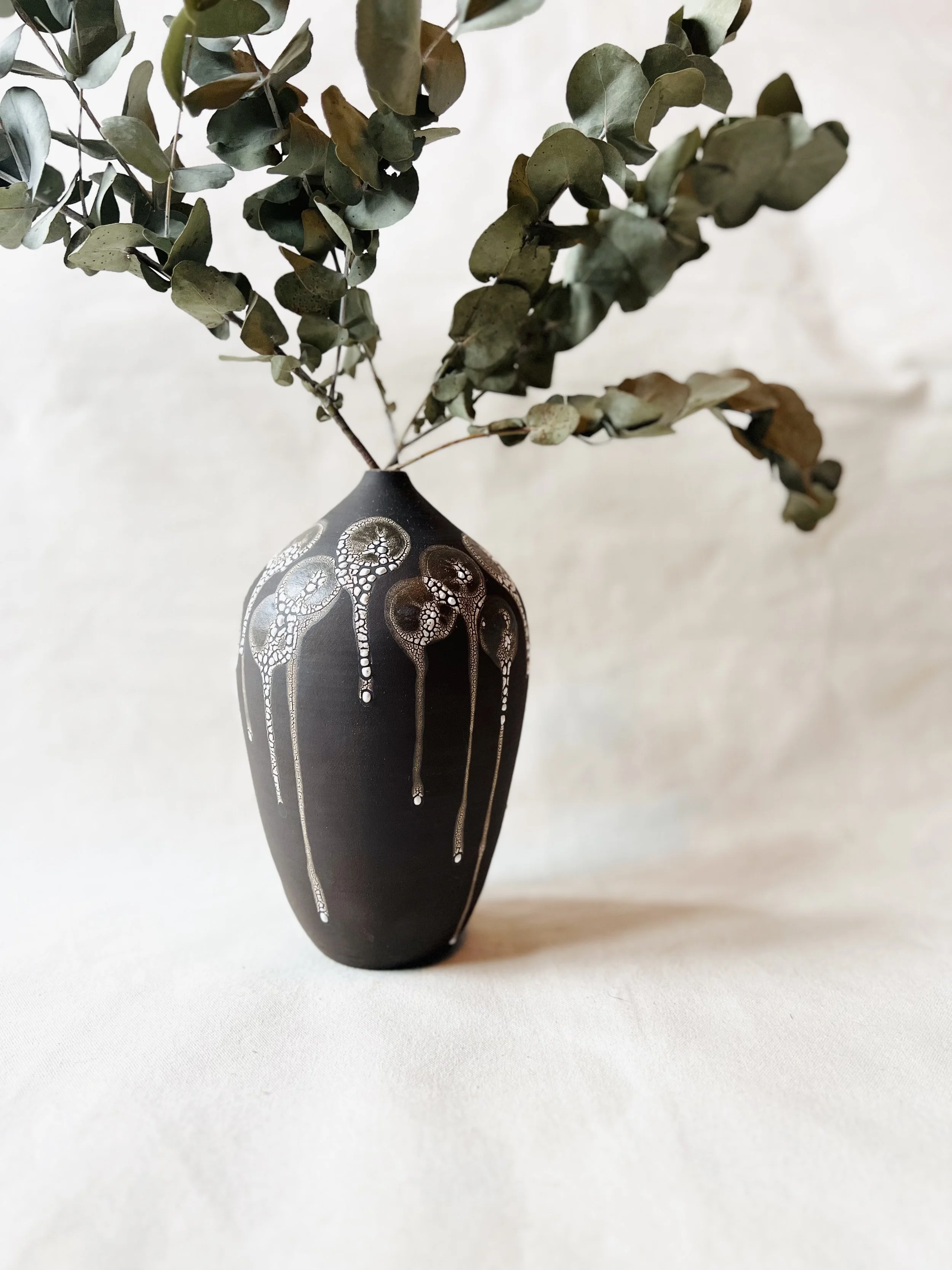 A black ceramic vase with white intricate patterns, holding green eucalyptus branches with oval leaves.