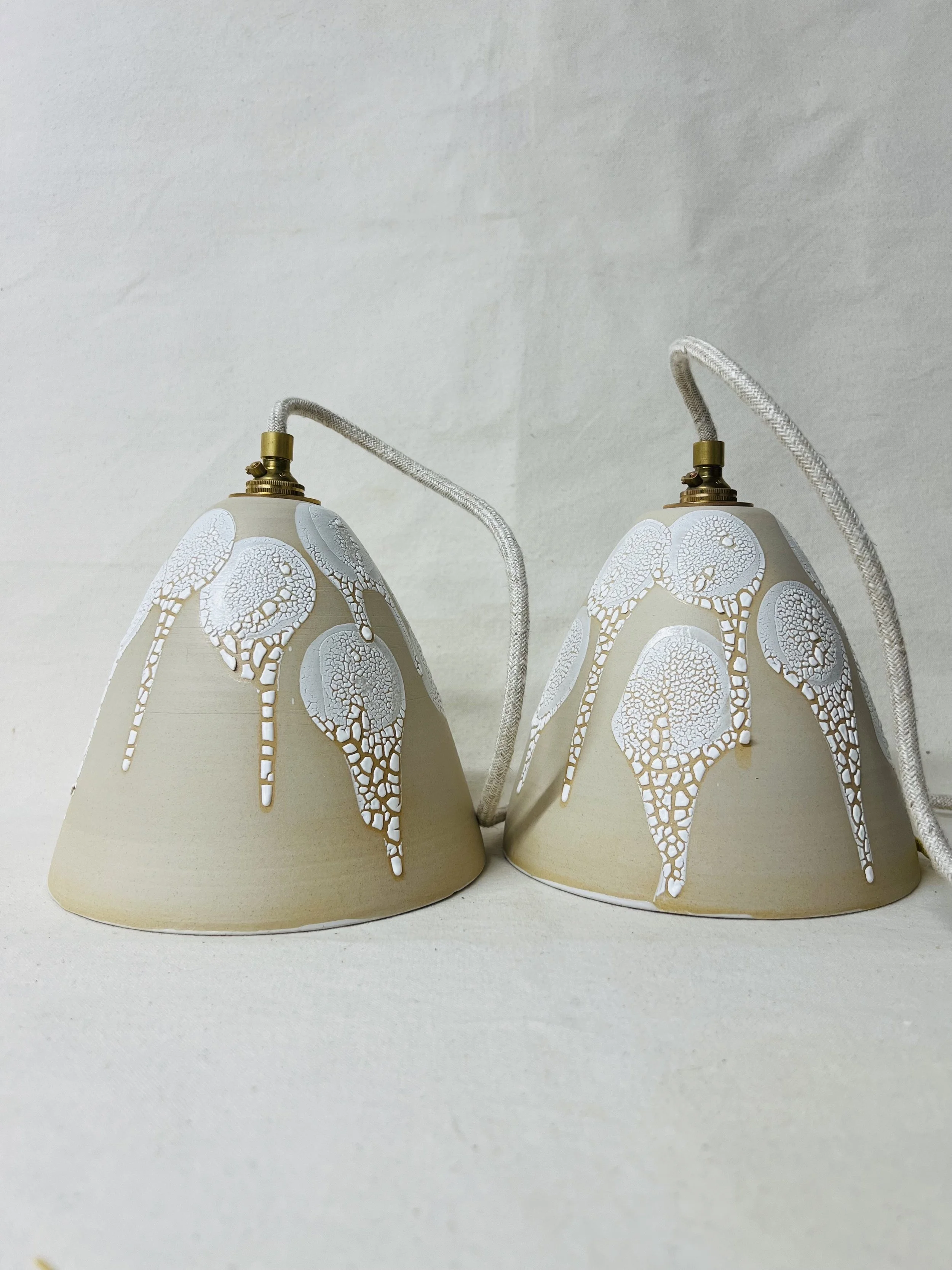 Two beige lamps with white lace-like patterns hanging from white cords against a white background.