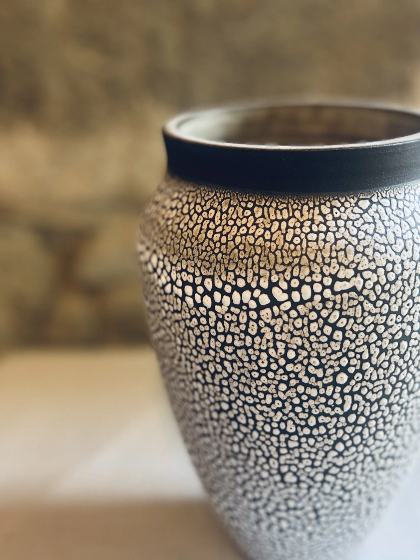 Close-up of a decorative vase with a black rim and a pattern of small white and black stones or beads on its surface, filled with a dark liquid, set on a white surface with a blurred background.