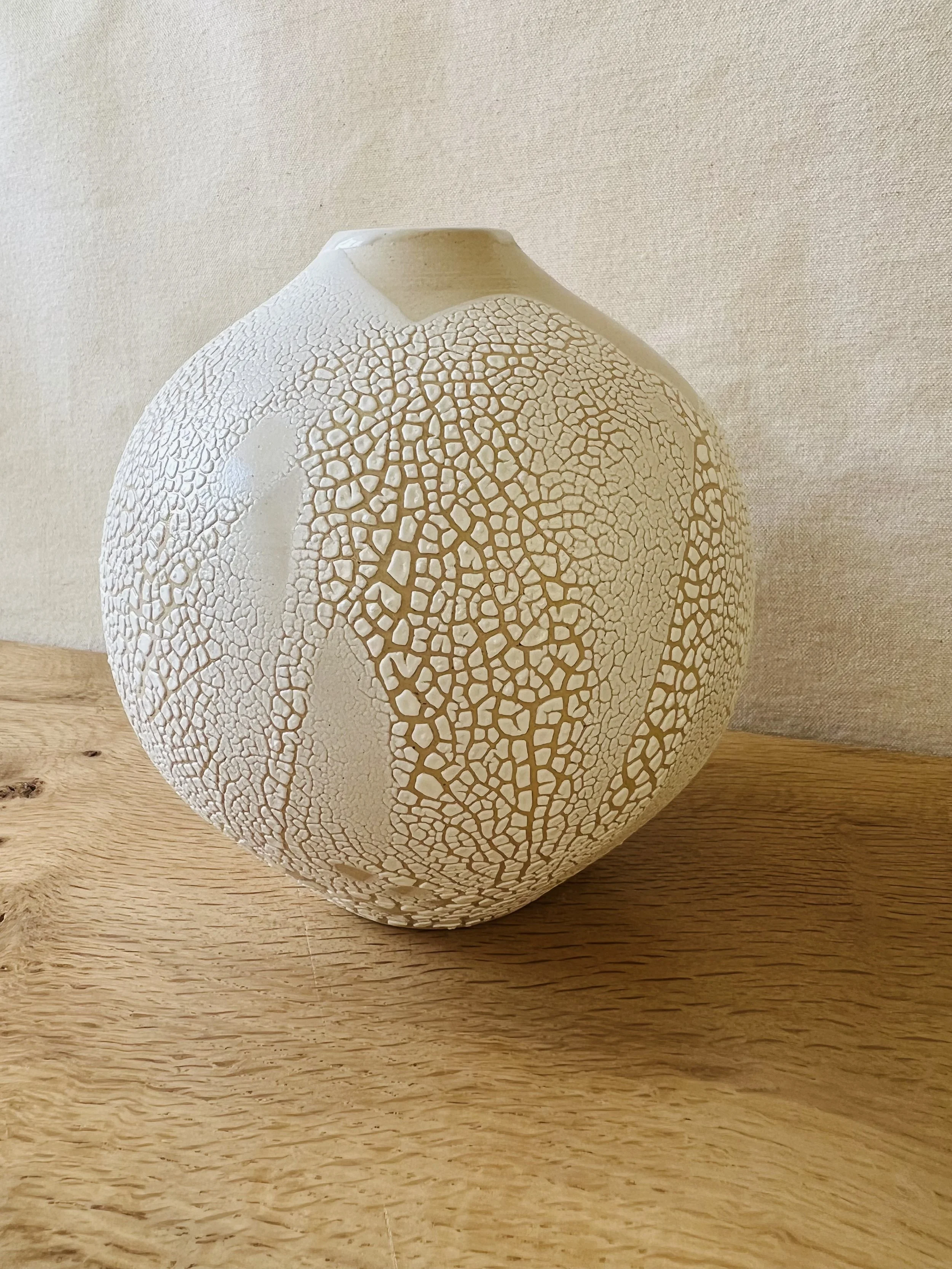A white textured ceramic vase with a cracked pattern, placed on a wooden surface against a beige fabric background.