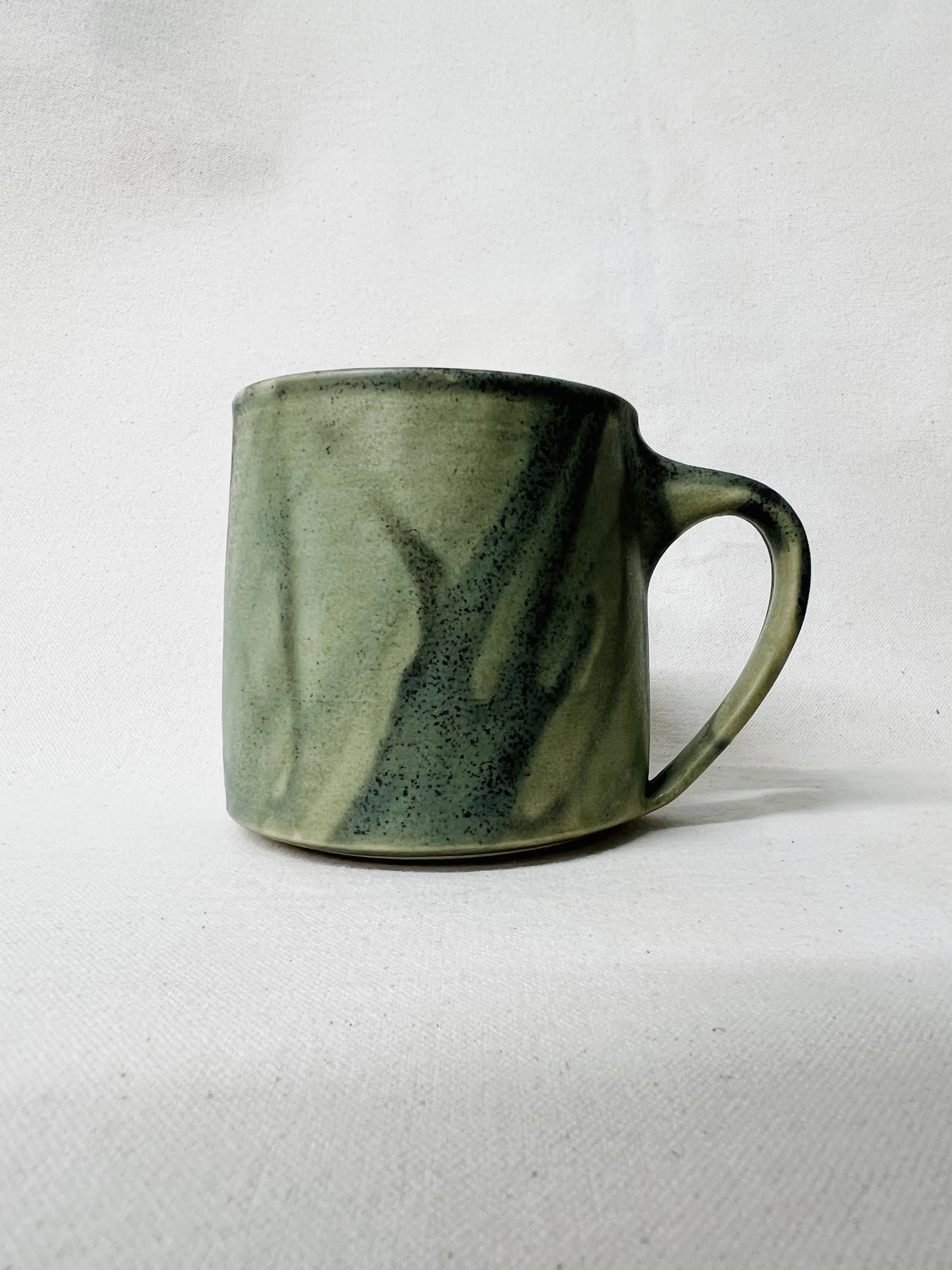 A ceramic mug with a marbled green glaze, placed on a white textured surface with a white background.