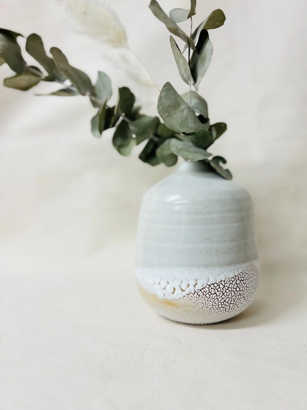 A minimalist ceramic vase with a textured pattern at the bottom, holding green leaves and branches against a neutral background.