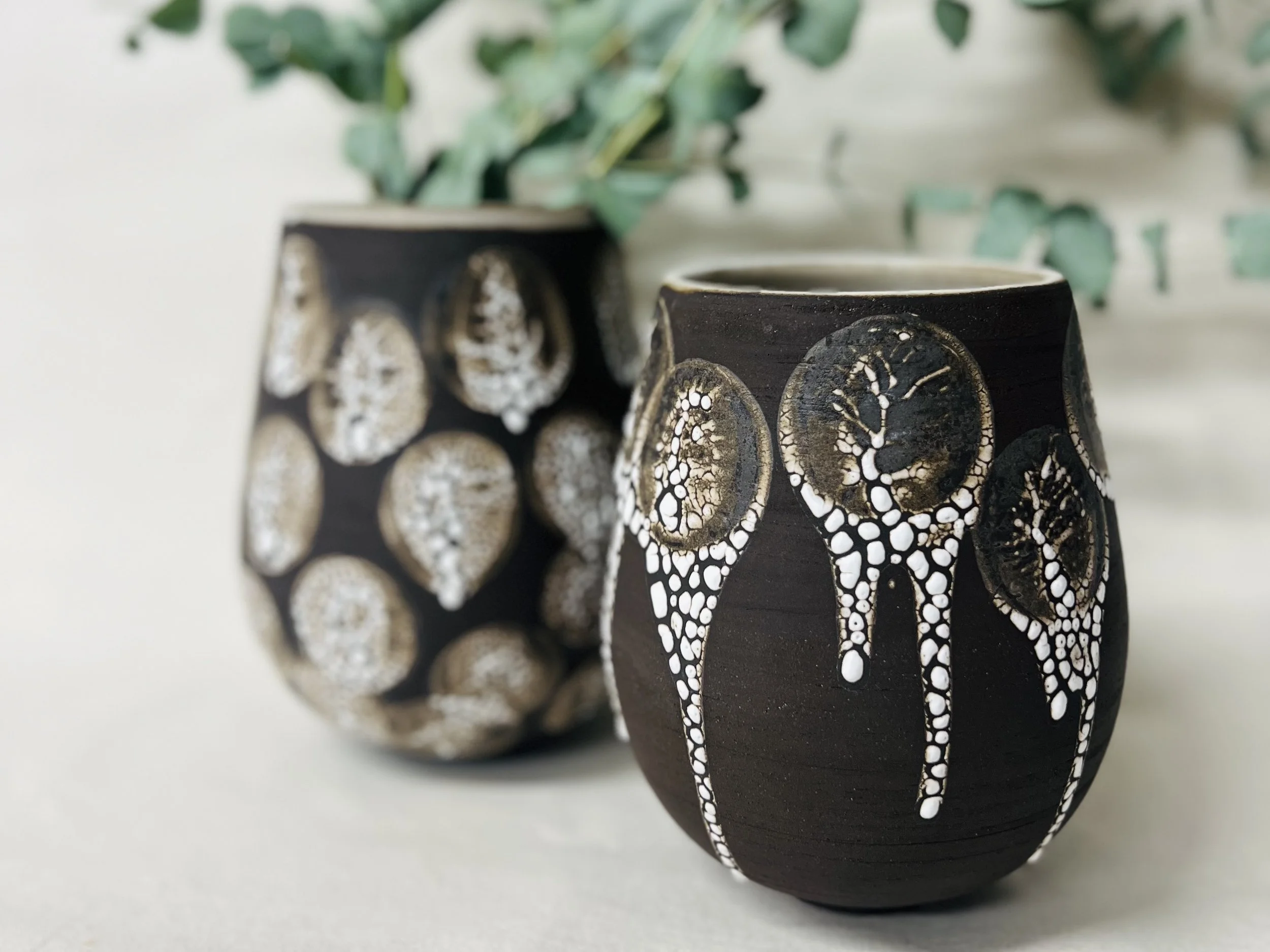 Close-up of two black ceramic vases with white and gold decorative patterns, and green plants in the background.