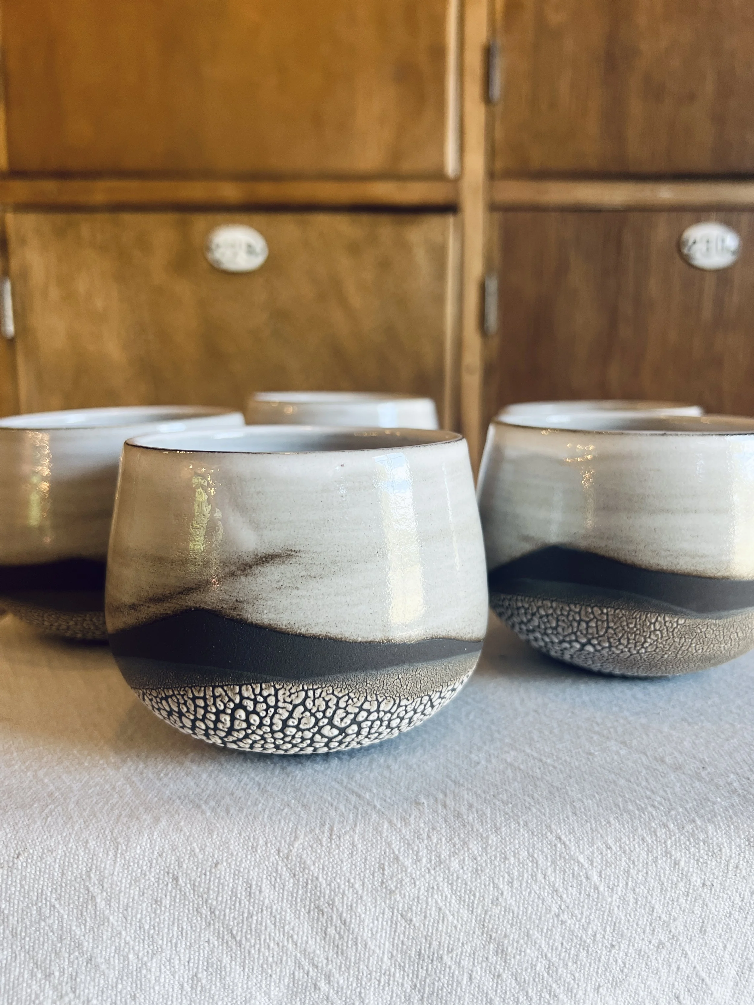 Three ceramic bowls with black textured bottoms and glaze patterns on the upper part, placed on a white fabric surface in front of wooden cabinets.
