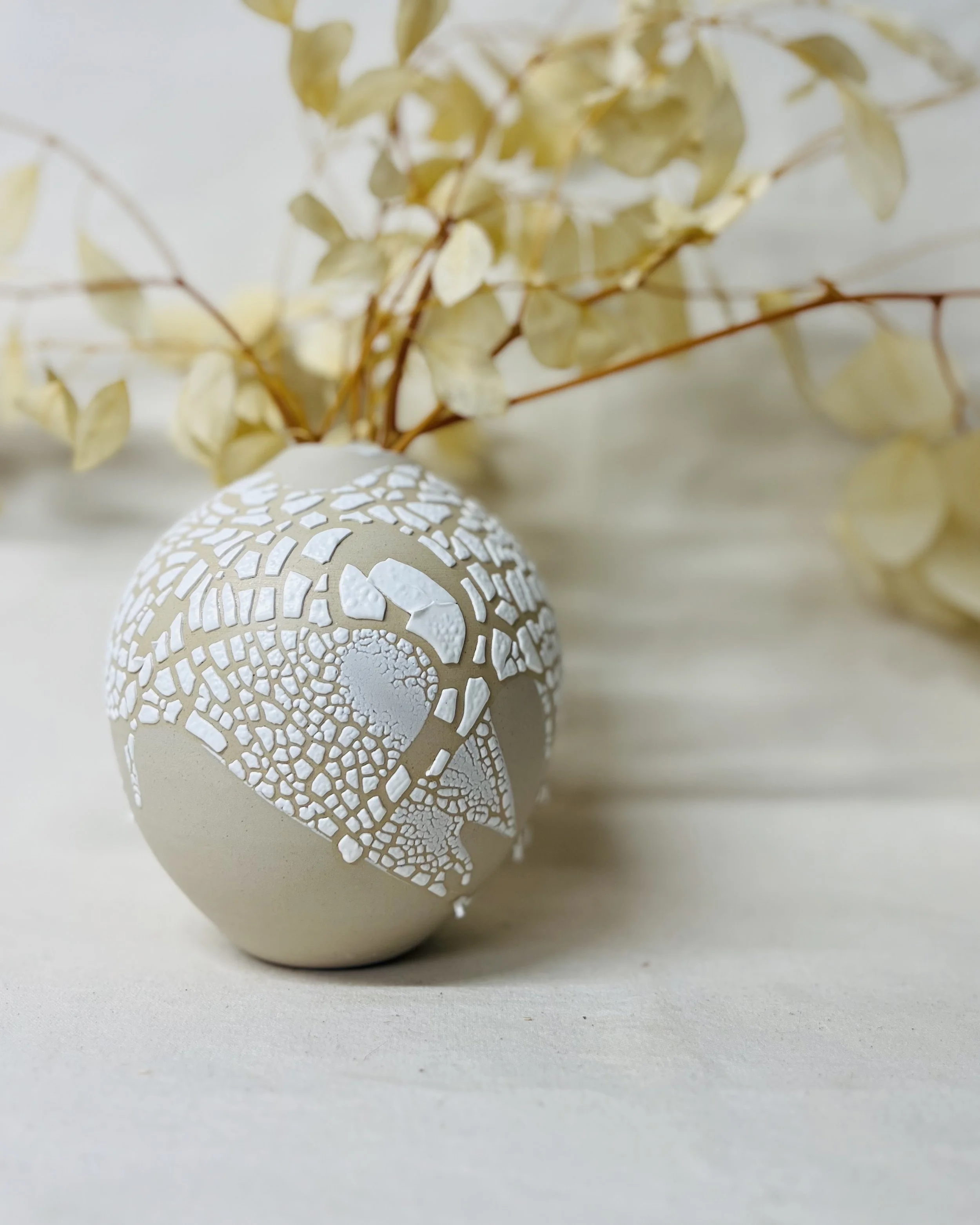 A white decorative egg with a pattern of intricate openings, placed on a light surface with a beige dried floral arrangement nearby.