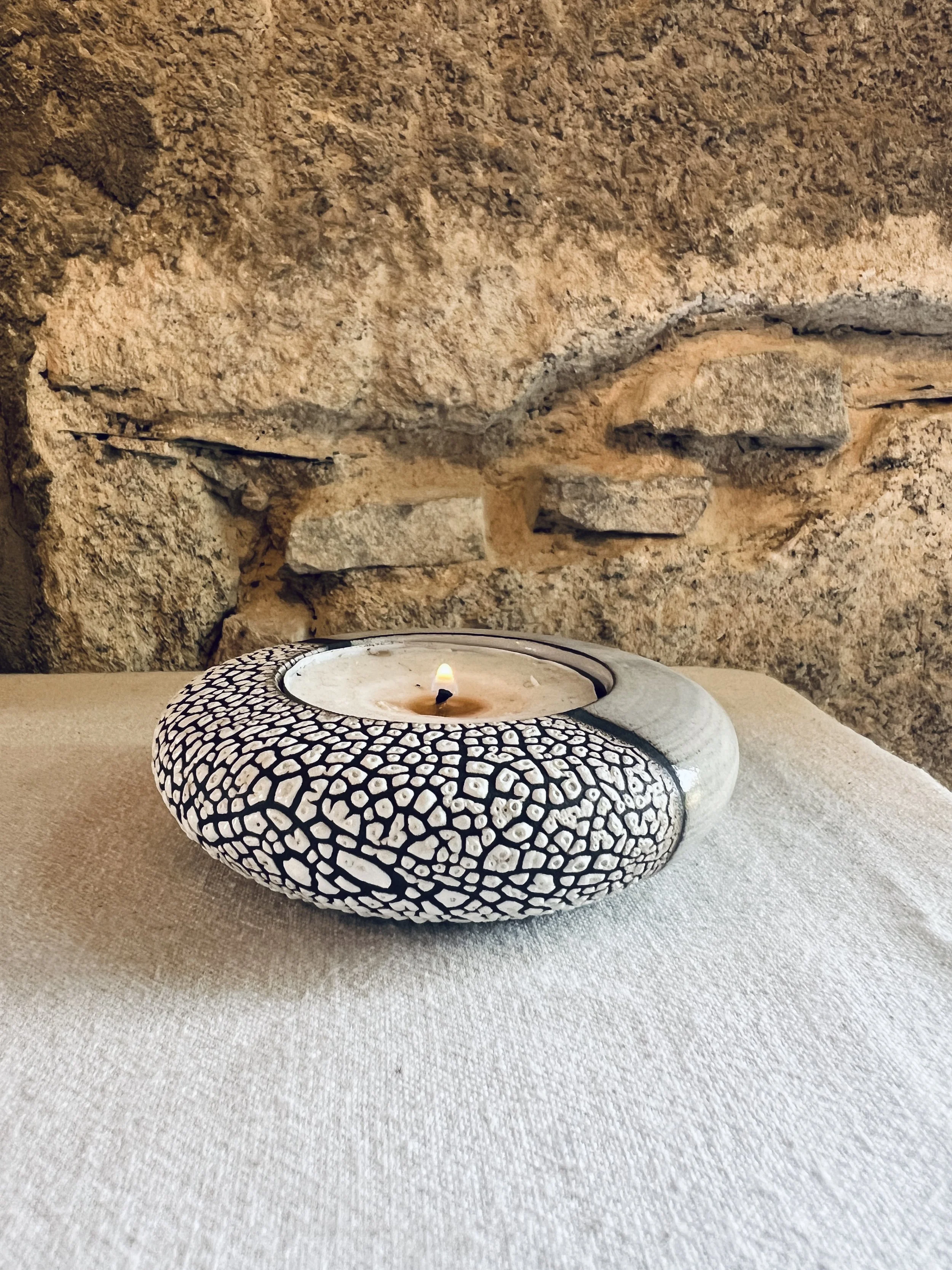 A lit candle in a decorative black and white patterned shell on a beige textured surface, with a stone wall in the background.