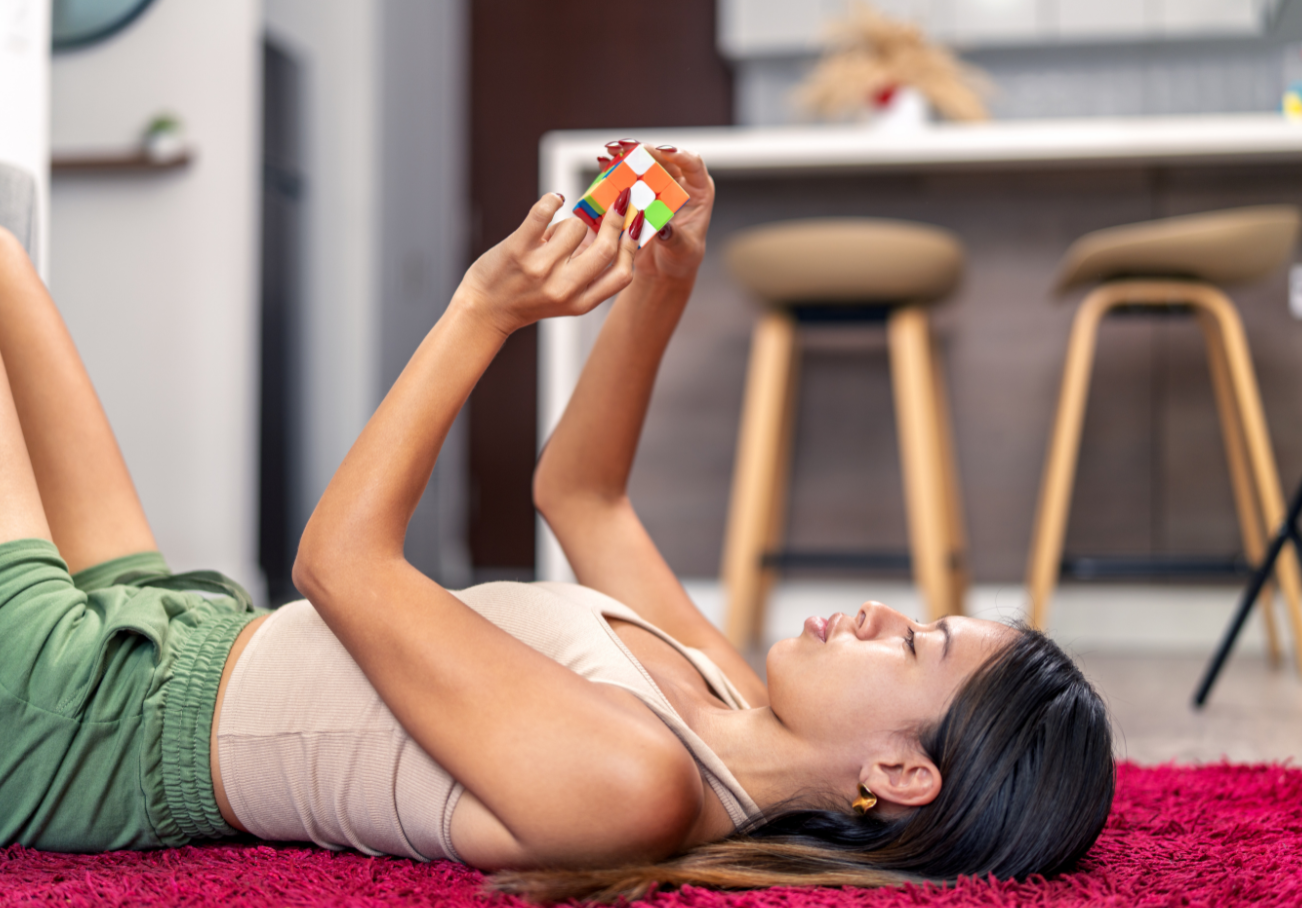 Woman lying on a pink rug, playing with a colorful Rubik's Cube in a modern living room.