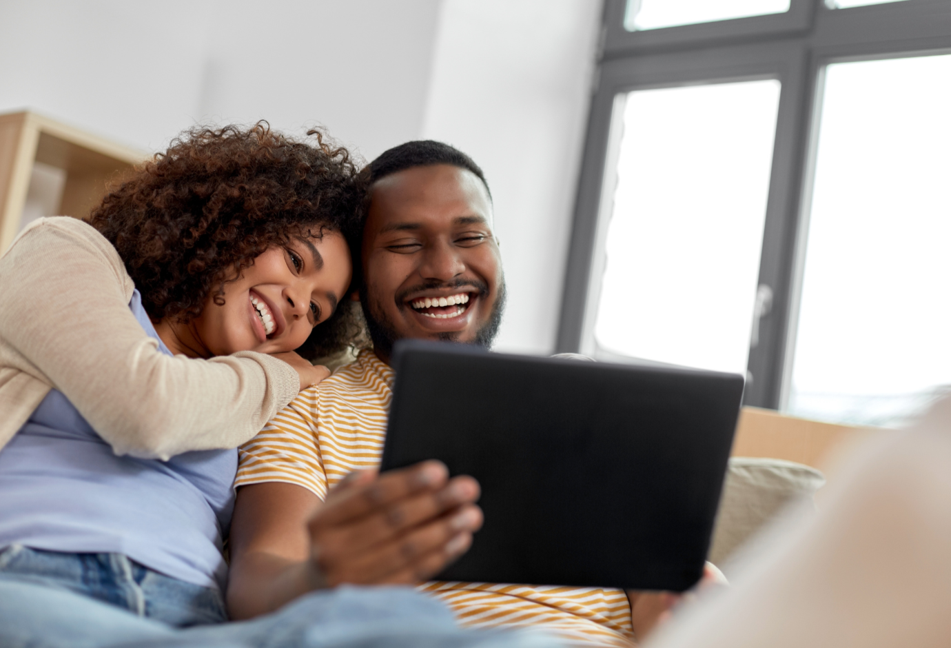 A happy couple sitting on a sofa, looking at a tablet and smiling.