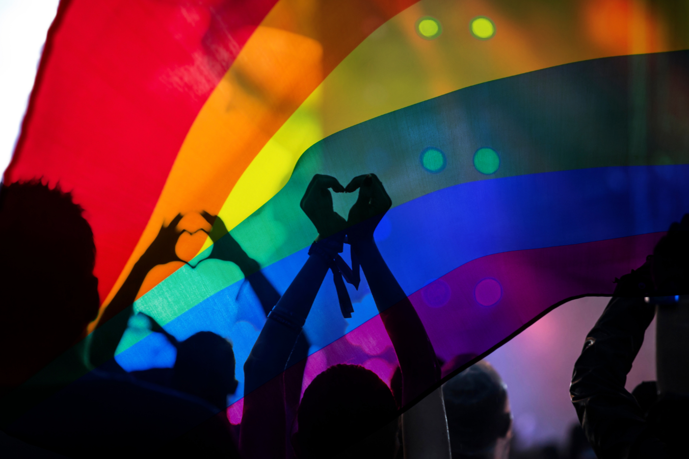 People holding a rainbow flag at a pride event, with silhouettes making heart shapes with their hands.