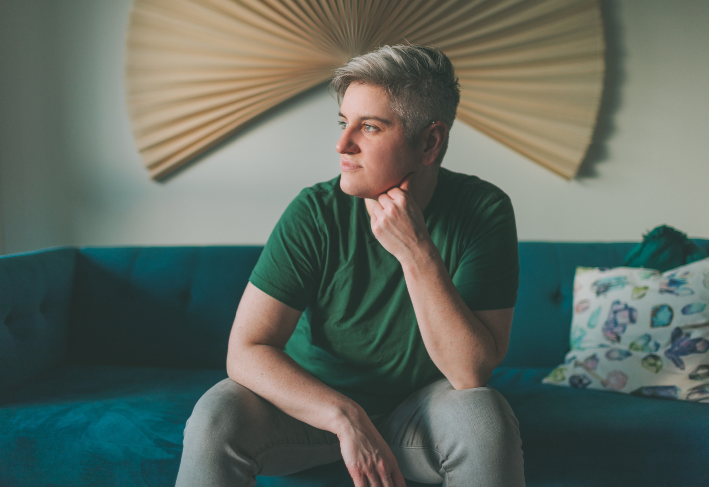 A young man with short gray hair, wearing a green t-shirt, sitting on a teal couch in a room with a large beige decorative fan on the wall behind him. He is resting his chin on his hand and looking to the side.