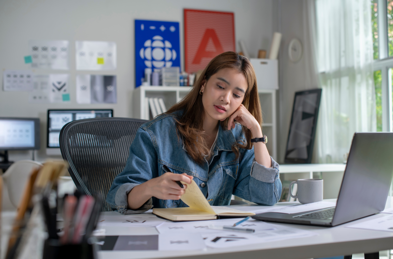 A young woman in a denim jacket working at a cluttered desk in a bright, modern office, reading a yellow note pad.