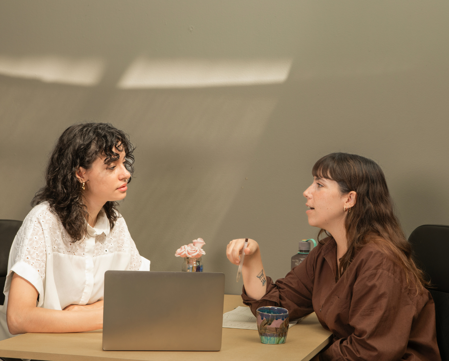 Two women sitting at a table, engaged in conversation, with a laptop and a cup on the table.