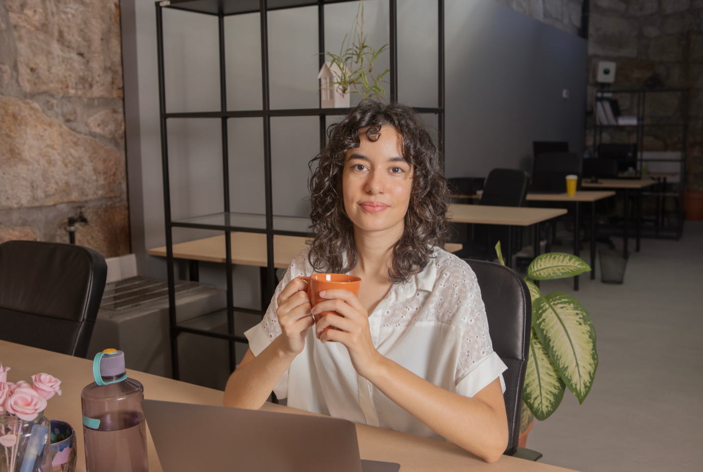 A woman with curly hair holding a mug and sitting at a desk with a laptop in an office.