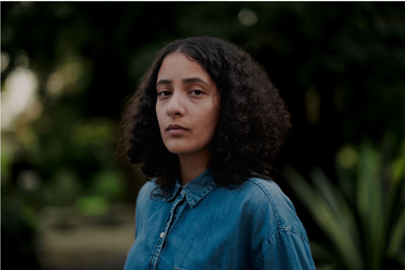 A young woman with curly black hair wearing a denim shirt, standing outdoors with a blurred green foliage background.
