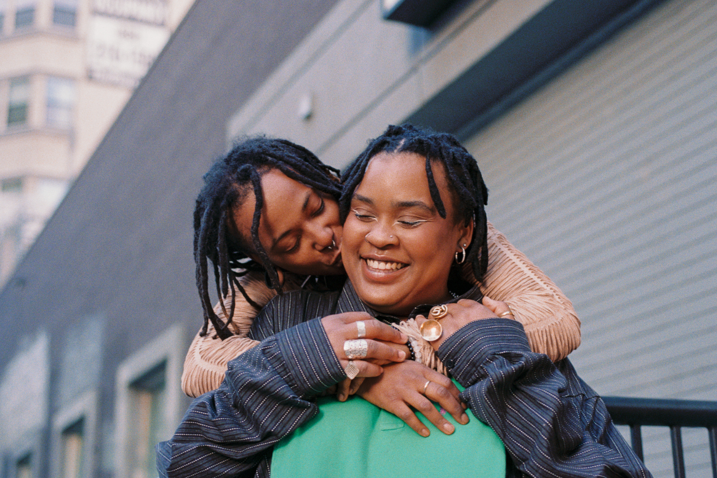 A woman with braided hair, wearing a striped jacket and jewelry, is smiling while being embraced from behind by a person with braided hair, wearing a tan jacket. They are outdoors in an urban area.