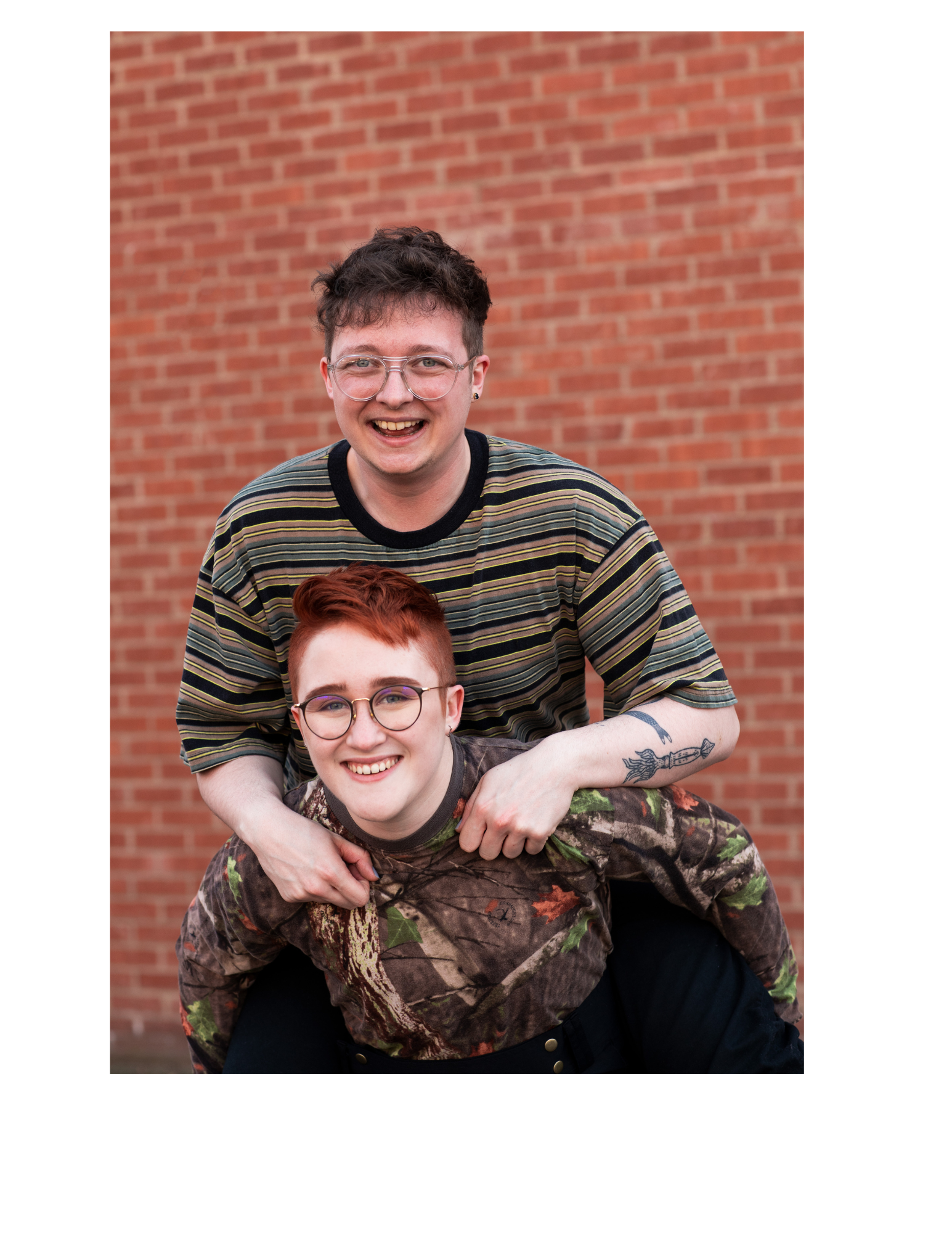 Two smiling young adults, one with short curly hair and glasses, and the other with short red hair, posing together in front of a brick wall.