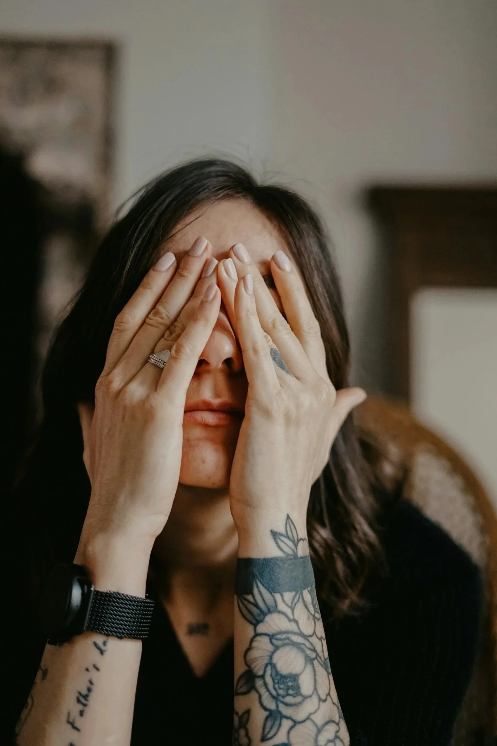 A woman with long dark hair covering her face with her hands, some tattoos visible on her arms, wearing a black watch and a ring, indoors with a blurred background.