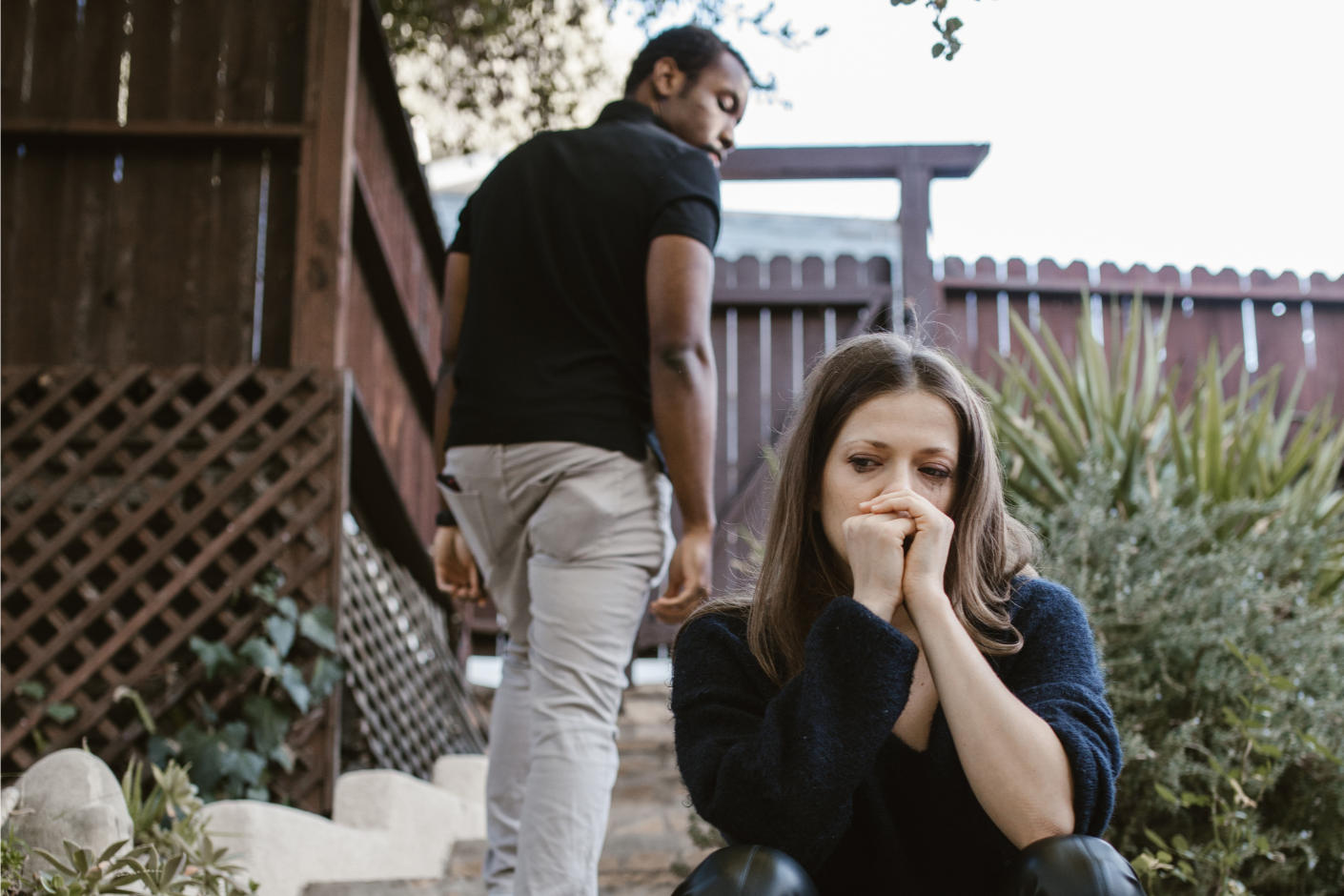 A woman sitting on the ground with a worried or anxious expression, covering her mouth with her hands, while a man stands behind her, looking away. They are outdoors in a yard with plants and a wooden fence.