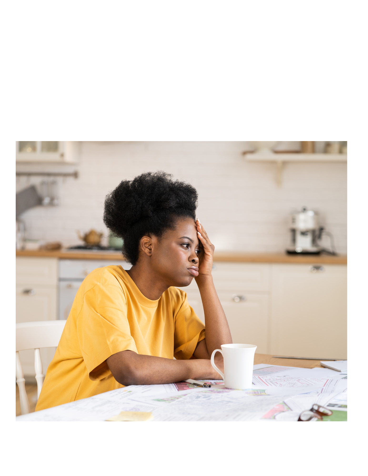 A woman sitting at a table with papers, a pen, and a white mug, appearing stressed or worried, with her hand on her forehead and a concerned facial expression in a bright kitchen.