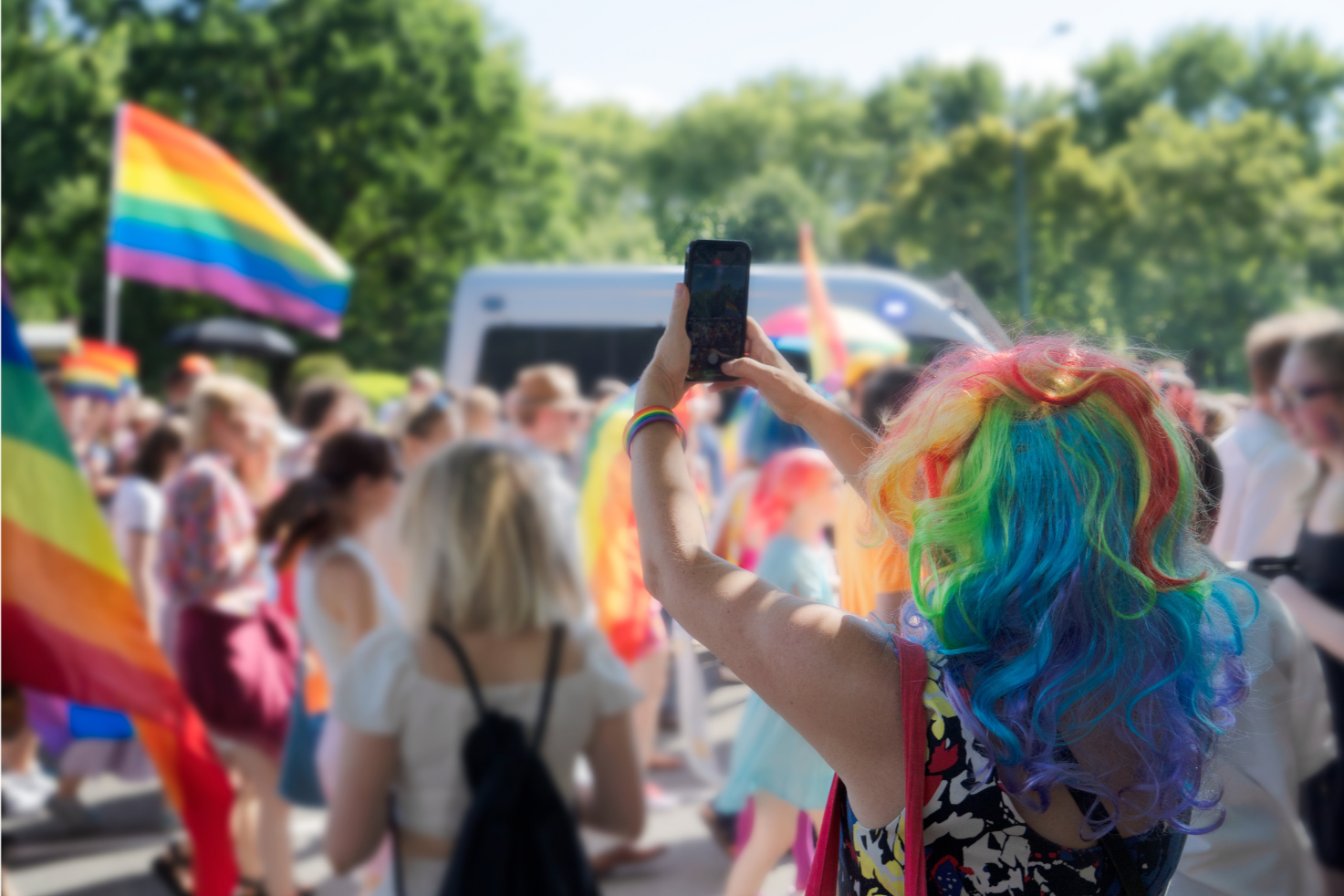 Person with rainbow-colored hair taking a photo at a pride parade, surrounded by a crowd of people, some holding rainbow flags, on a sunny day with green trees in the background.
