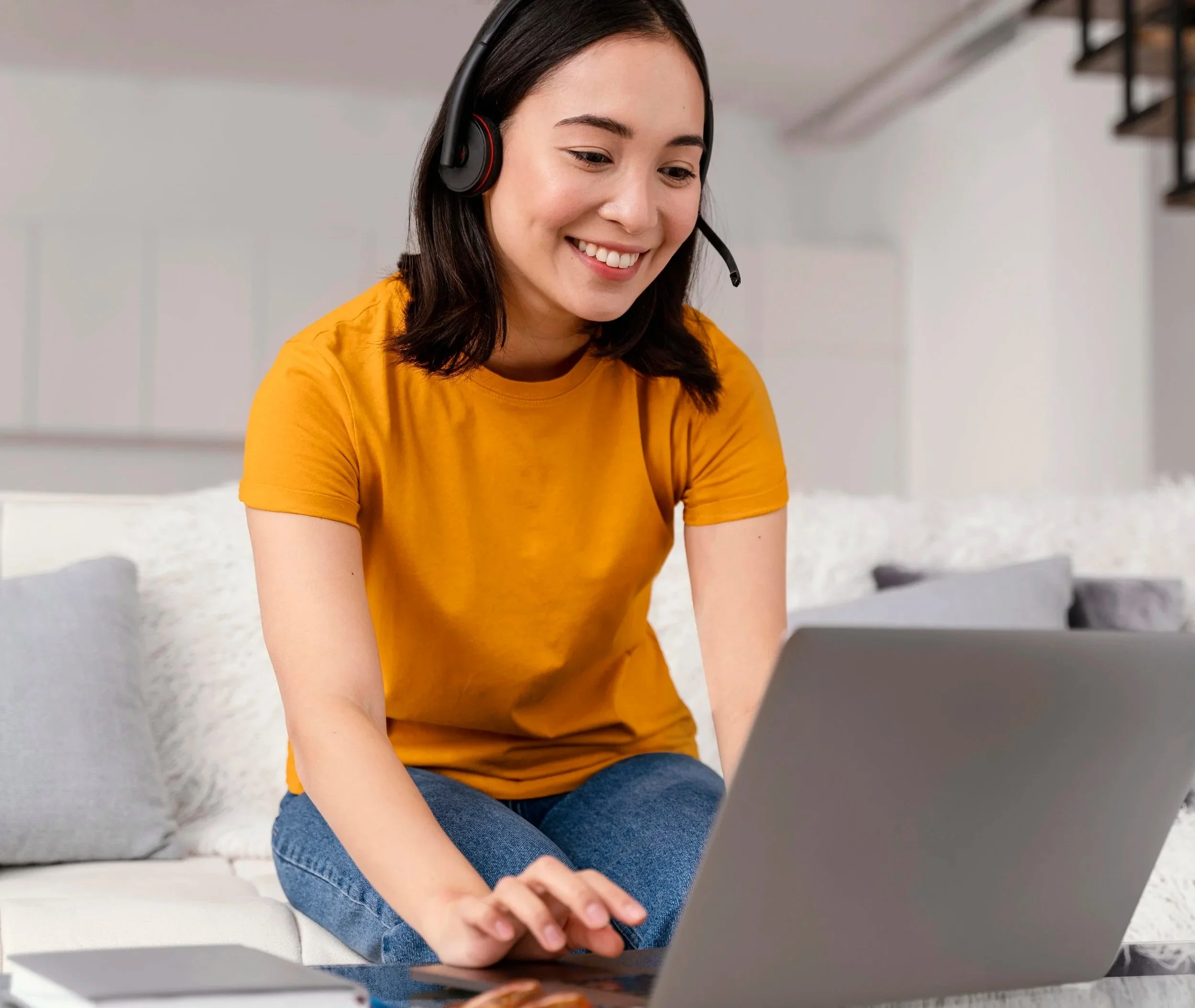 A young woman with dark hair wearing an orange shirt and jeans using a headset, sitting on a white bed with gray pillows, working on a laptop in a bright room.