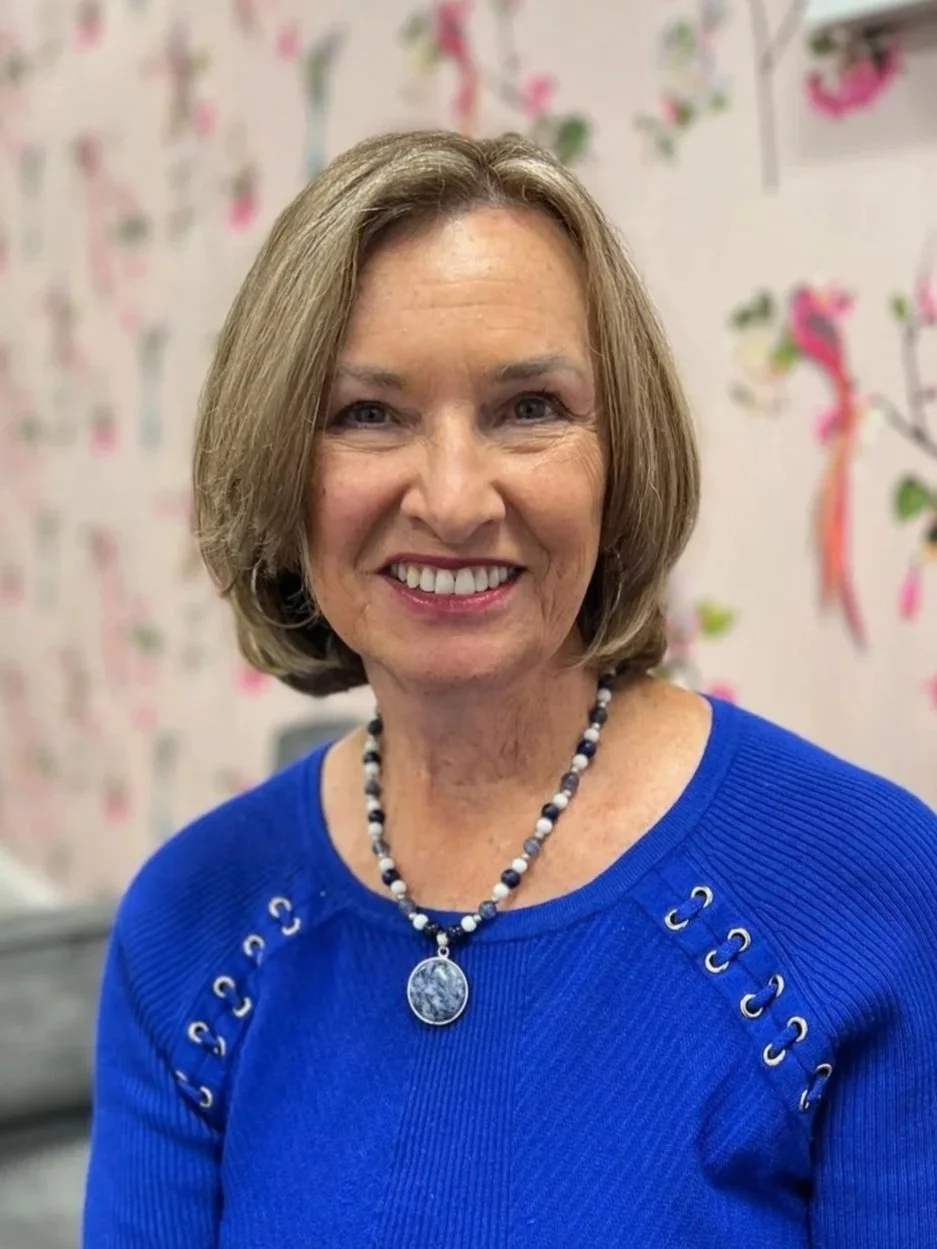 A smiling woman with short blonde hair wearing a bright blue sweater and a beaded necklace, sitting in a room with a pink wall decorated with colorful bird and flower illustrations.
