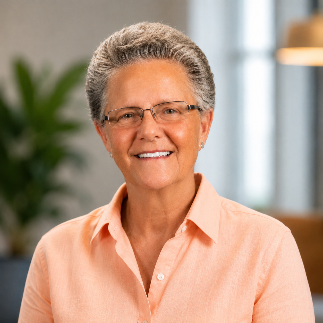 A woman with short gray hair wearing glasses and a pink collared shirt, smiling indoors.