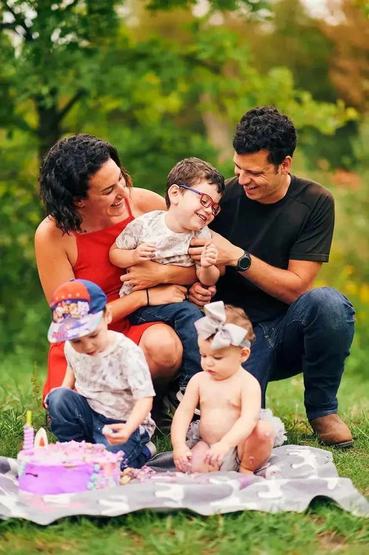 A family celebrating a child's birthday outdoors with a cake and children, including one with a large gray bow, in a park.