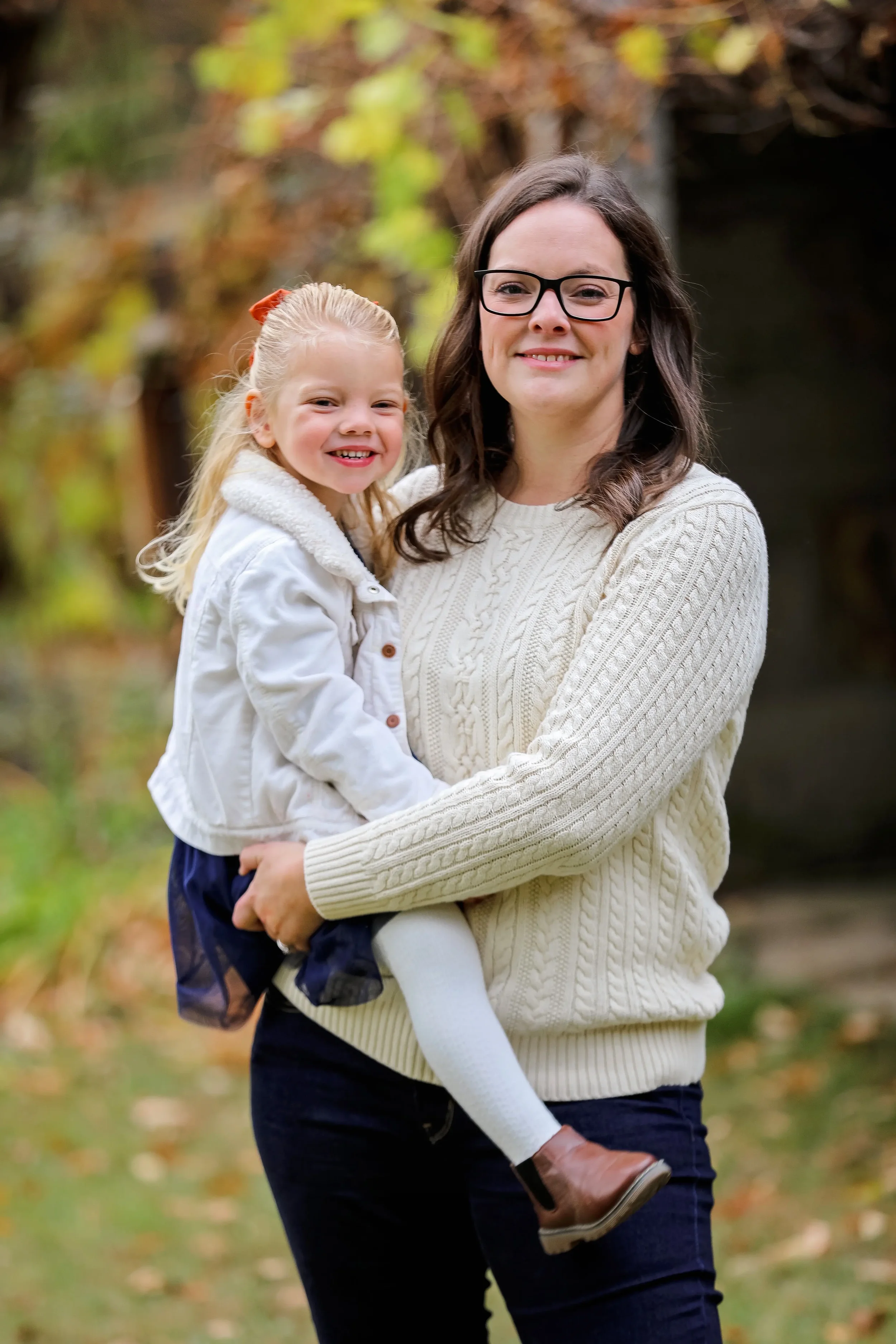 A woman with glasses and long brown hair smiling while holding a young girl with blonde hair and a white jacket outdoors in autumn.