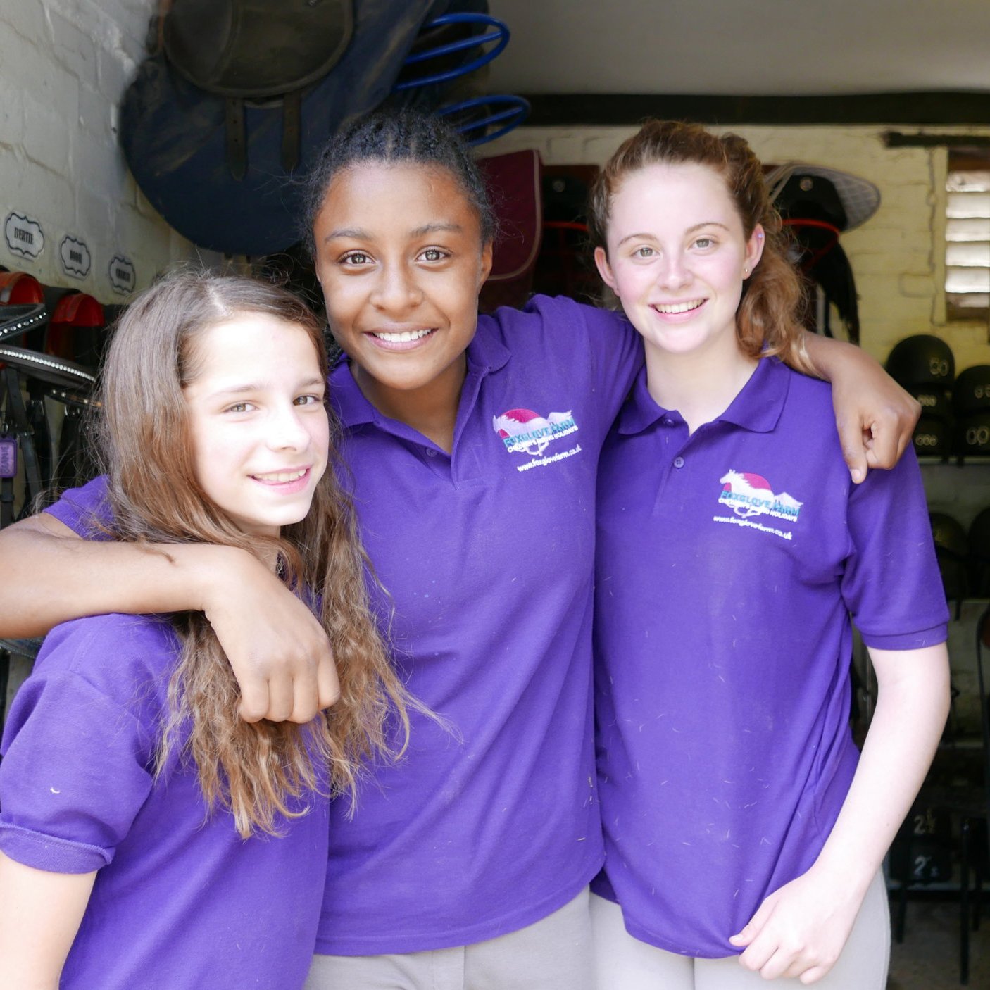 Three girls in a horse stable looking at the camera and smiling with arms around each other wearing Foxglove Farm t-shirts in signature purple with logo designed by Yeswaydesign
