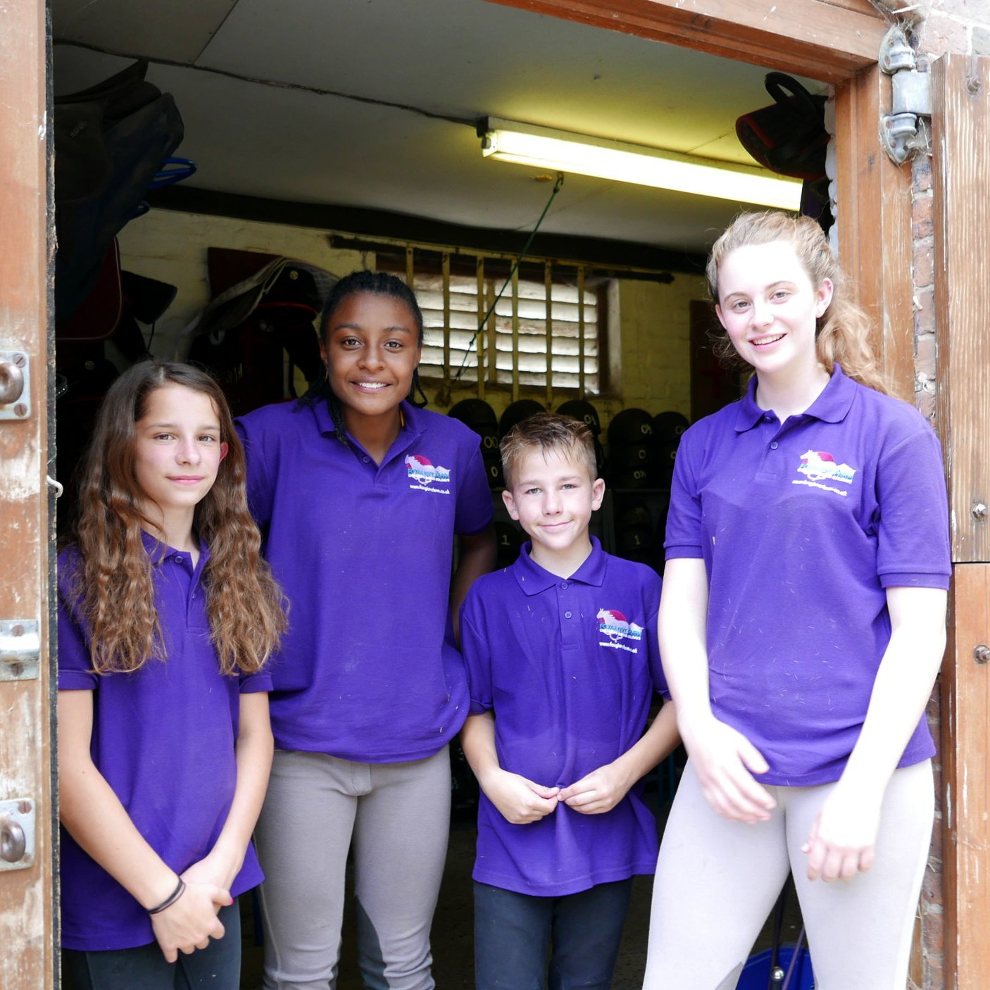 Three young girls and a boy in a horse stable looking at the camera and smiling wearing Foxglove Farm t-shirts in signature purple with logo designed by Yeswaydesign