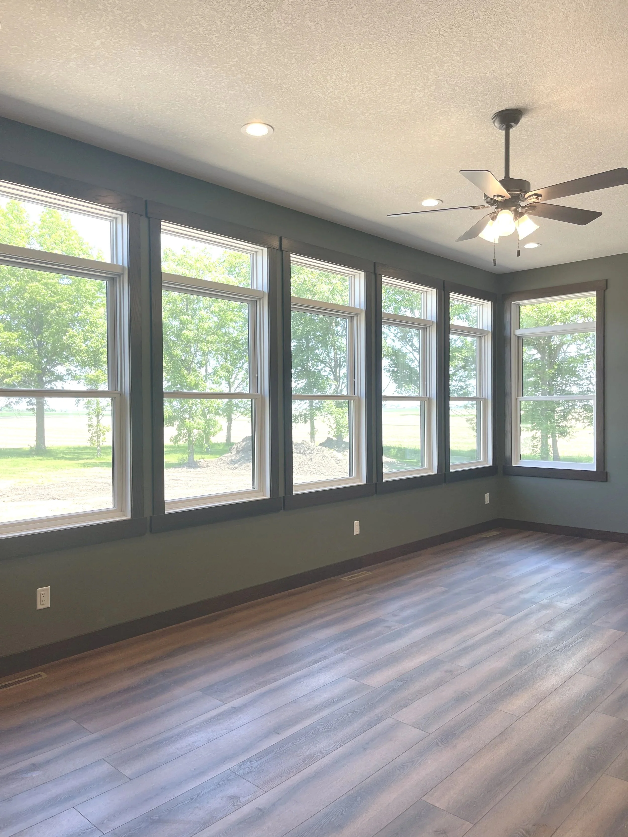 Empty room with large windows, ceiling fan, hardwood floors, and view of green trees outside.