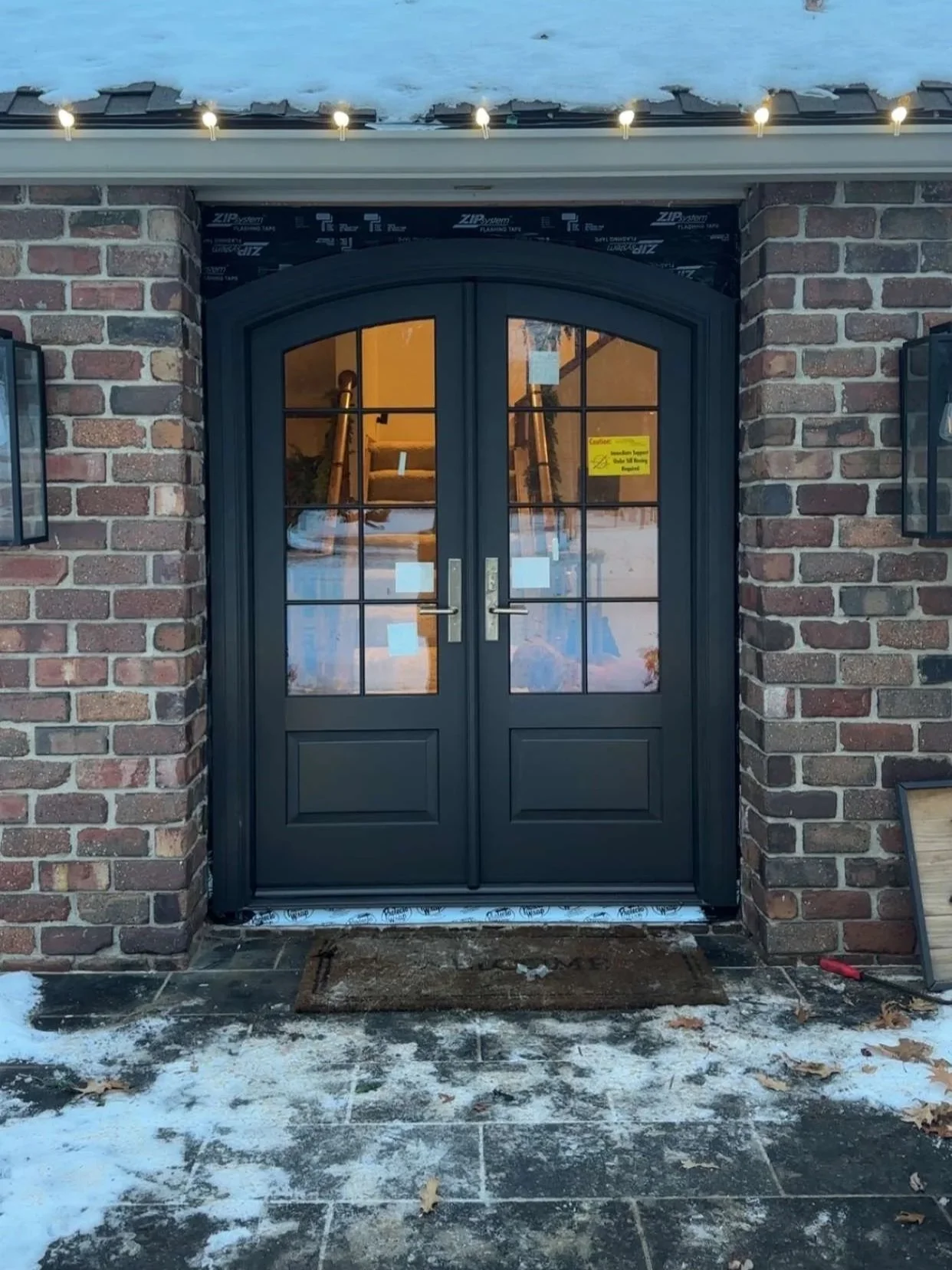 Backyard view of a house with white siding, large windows, and a sliding glass door. There is a concrete patio, a small garden bed with rocks and a few plants, and a white privacy fence. Overcast skies are visible.